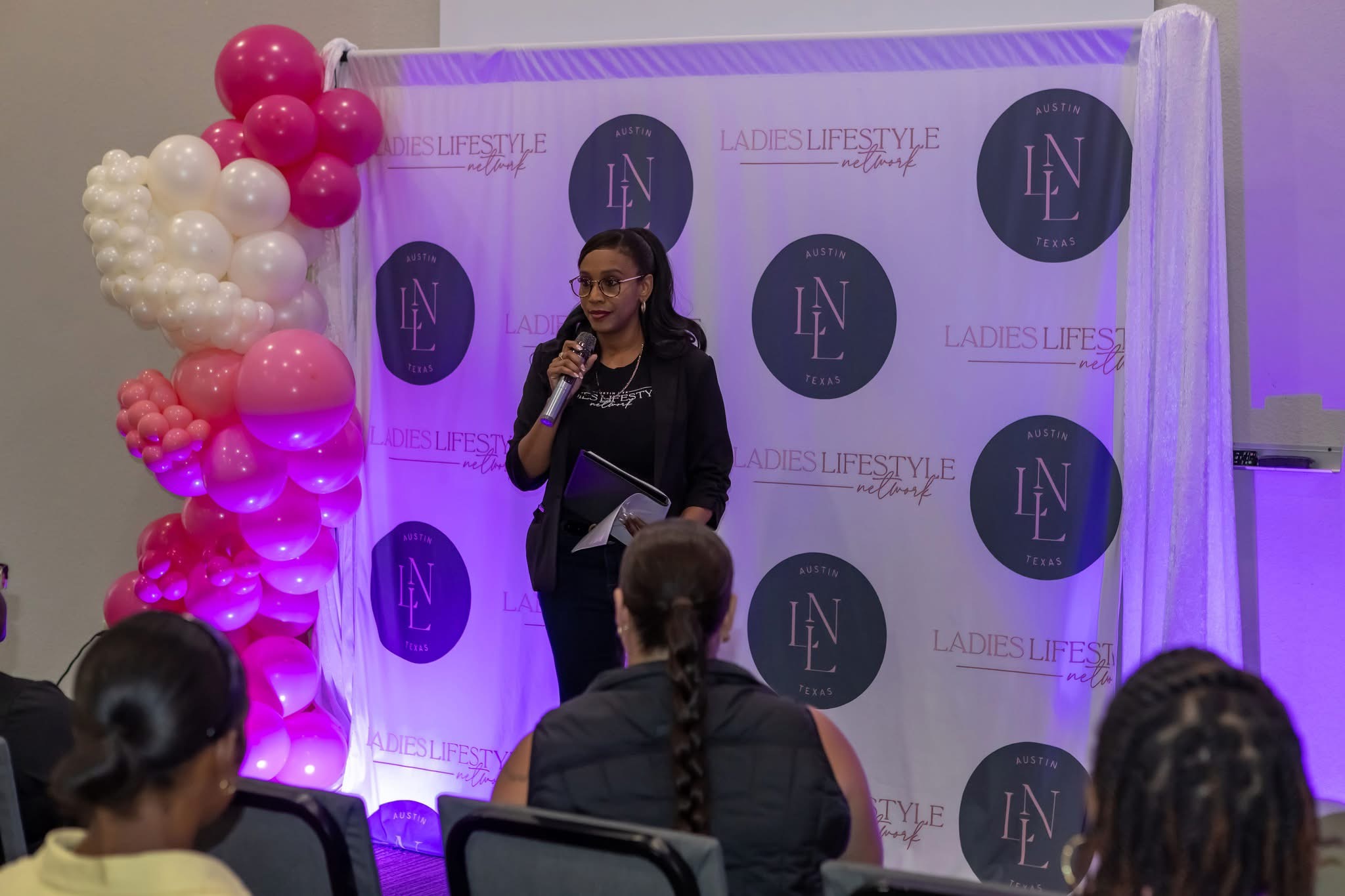 Woman speaking into microphone on stage with backdrop and balloon decoration, audience seated in front.