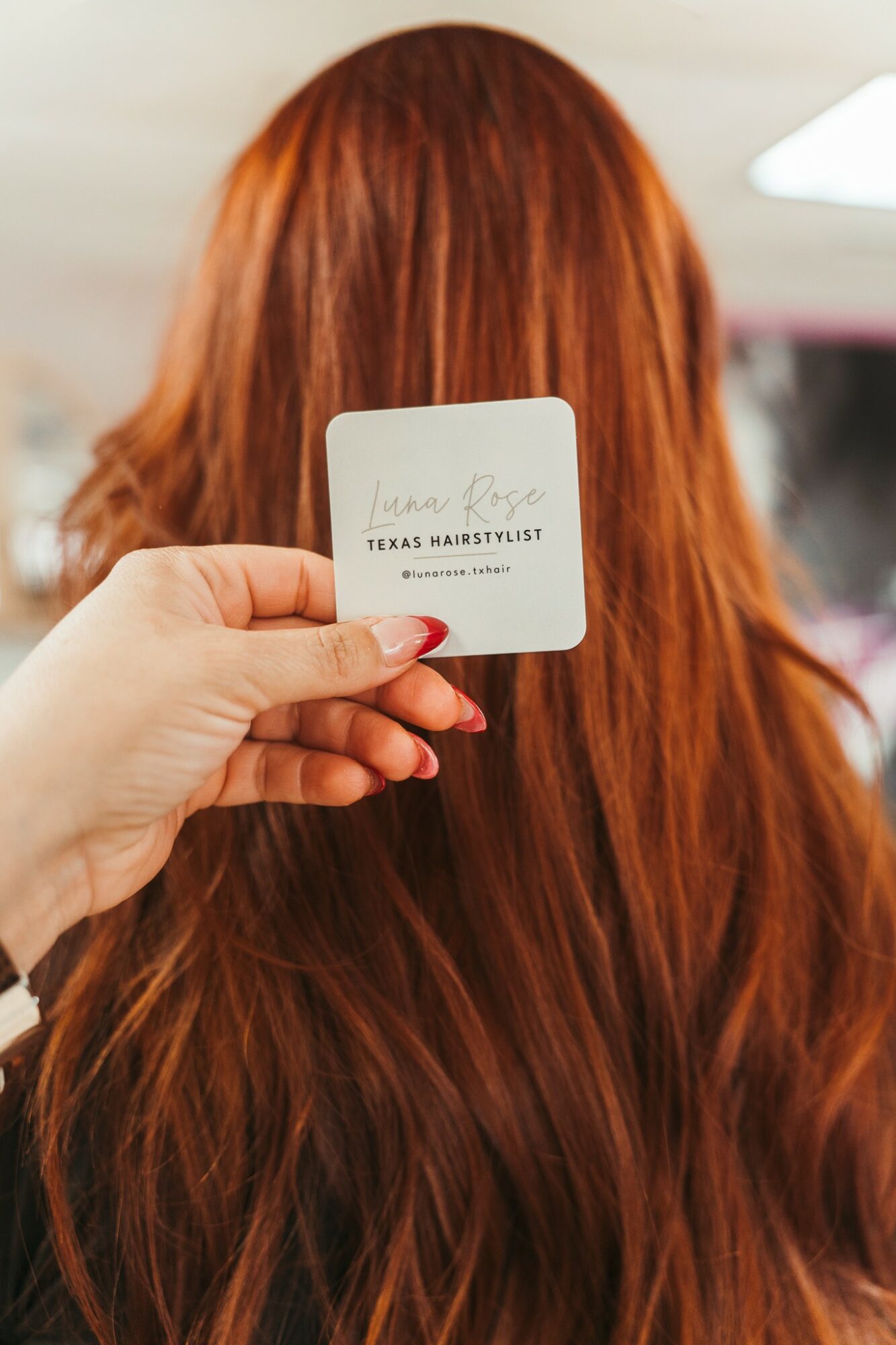 Hand holding a card in front of a person with long, red hair, indoors with ceiling lights visible