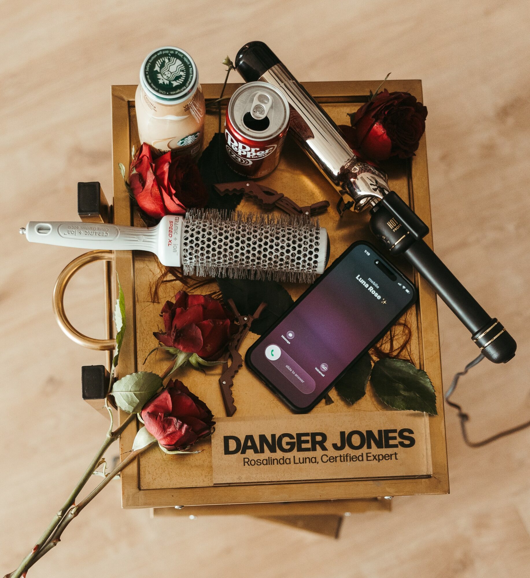 A wooden box contains a flashlight, a smartphone, a can, a small bottle, a key, and dried roses, on a wooden surface.