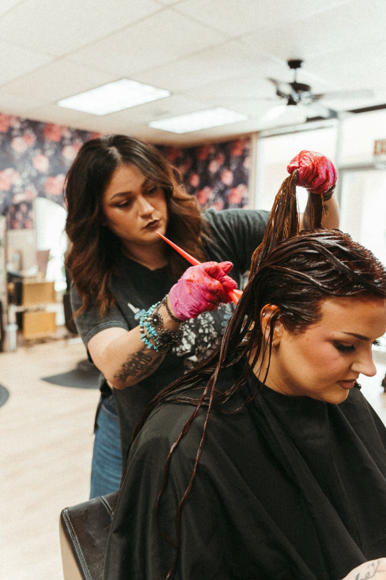 Hairdresser applying hair dye to a client's wet hair in a salon, with floral wallpaper in the background.