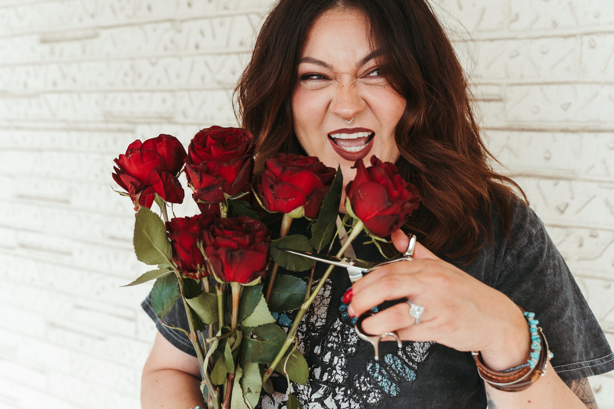 Woman with dark hair holding a bouquet of red roses, making a playful face, standing against a white brick wall.
