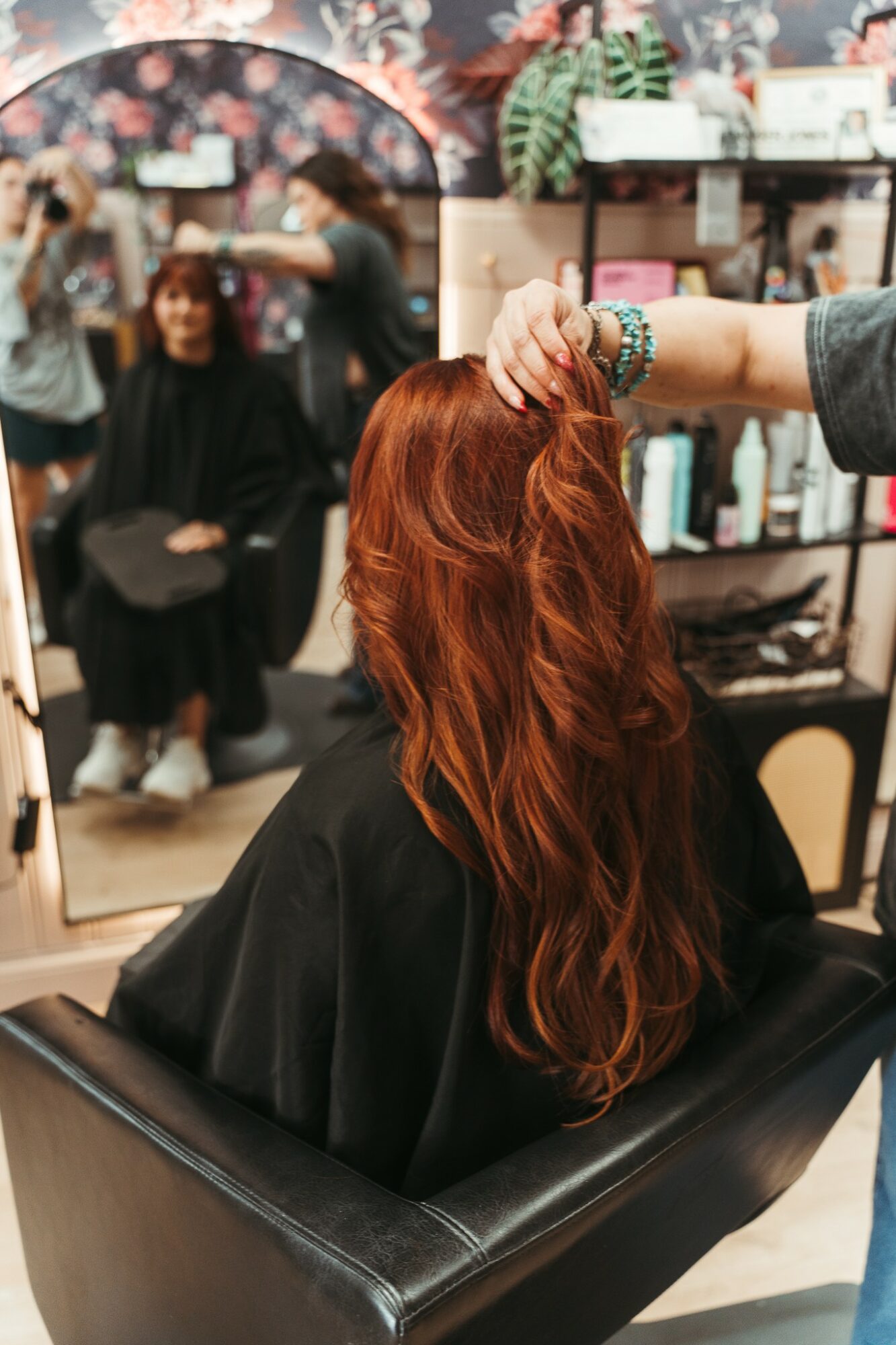 Person with long red hair sitting in a salon chair, hair being styled, mirror and shelves in background.