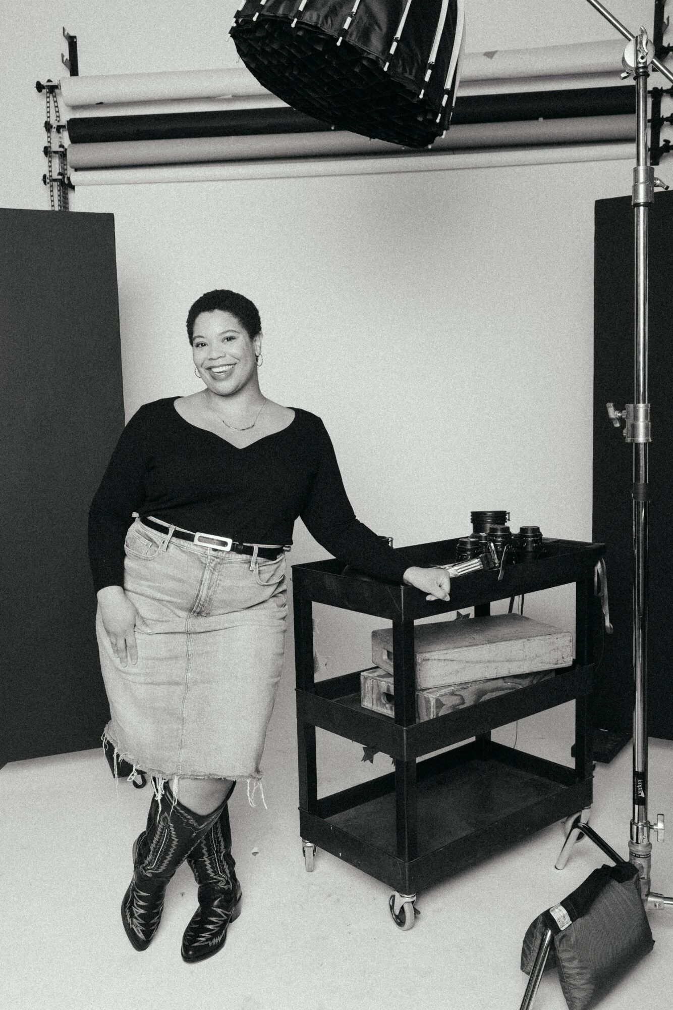 Woman smiling, standing next to a cart with photography equipment in a studio, black and white photo.