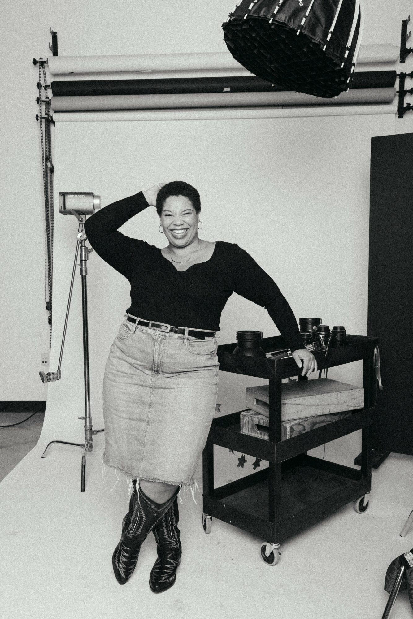 Woman smiling, standing with hand on head, in a photography studio with equipment, black top, light skirt, and boots.