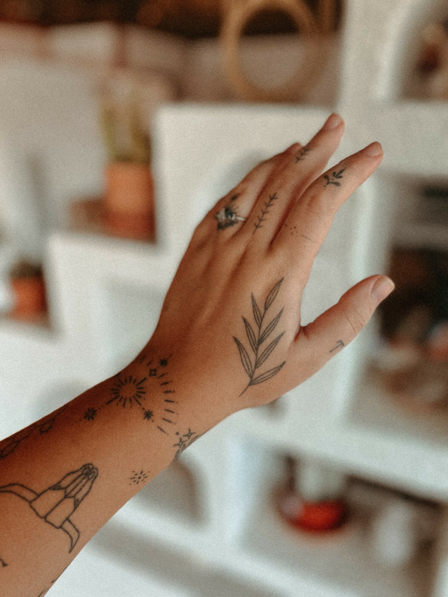 Hand with tattoos, including a leaf design on the back, a sun, and a figure, against a blurred background.