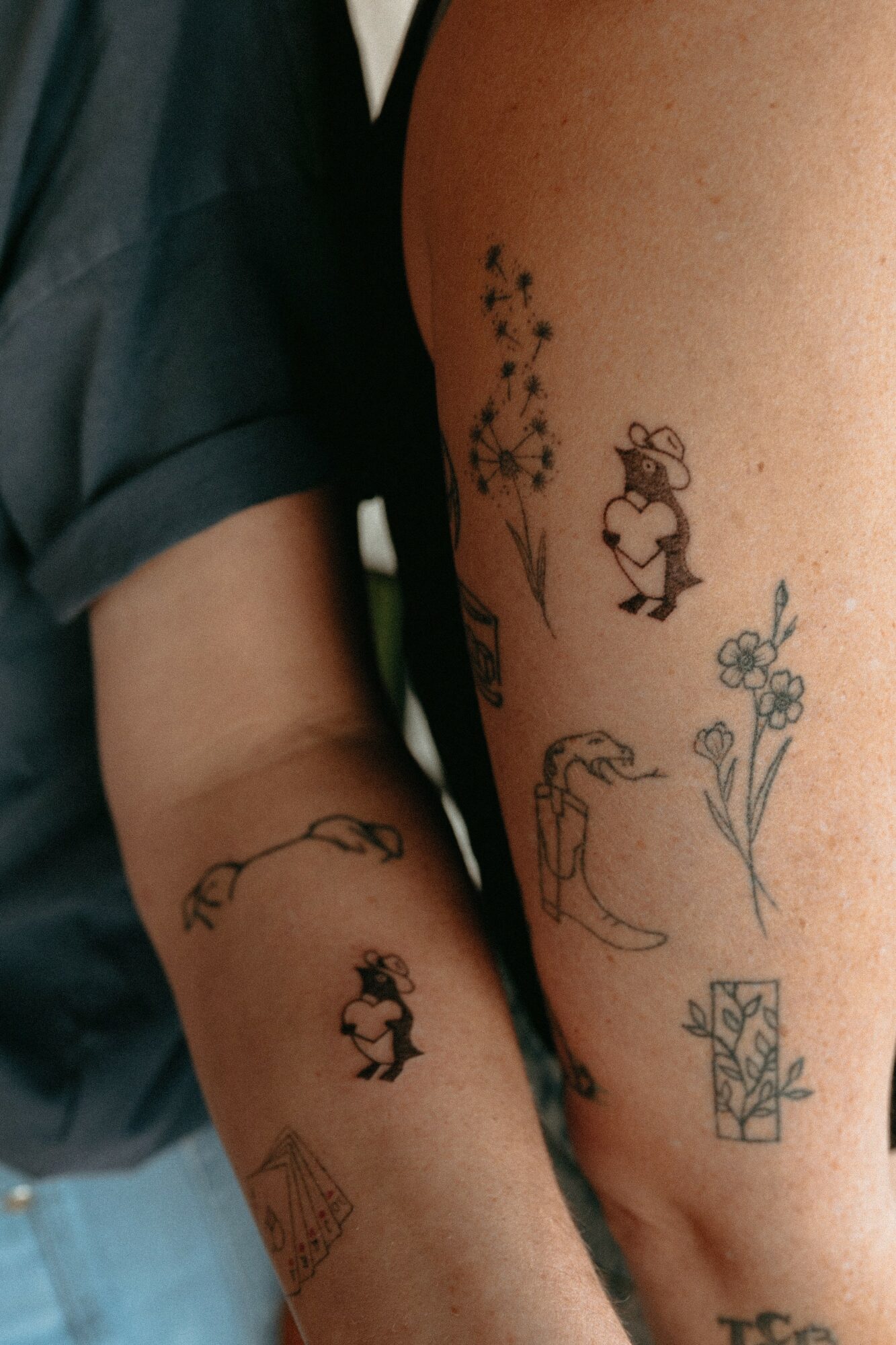 Two arms with various small tattoos, including a girl, flowers, a gift box, and a bird, close together.