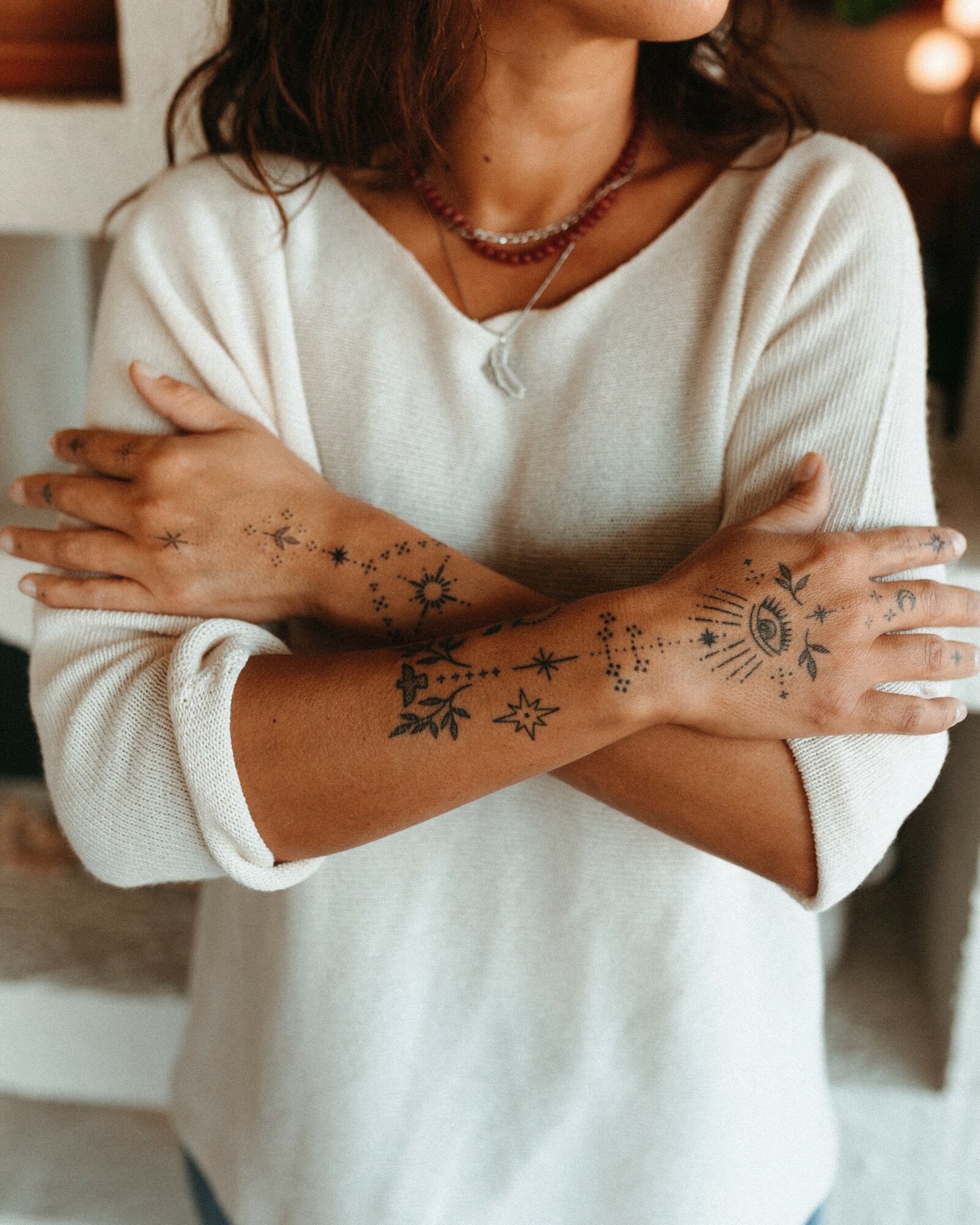 Person with tattoos on arms, wearing a white sweater and layered necklaces, crossing arms indoors.