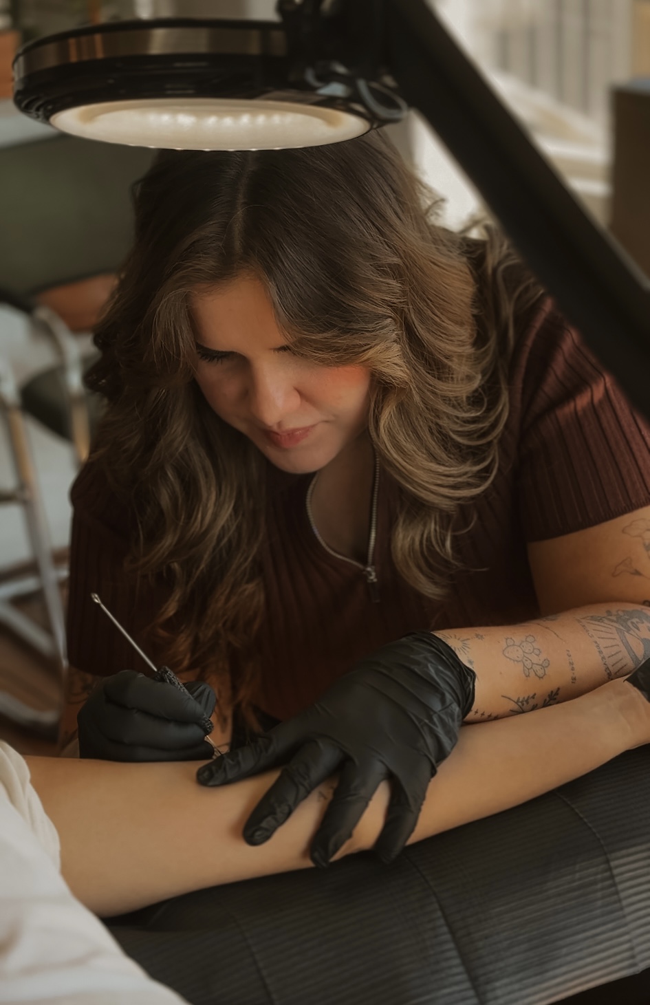 Woman with long wavy hair, wearing black gloves, tattooed arm, focused on tattooing someone's arm under a lamp.