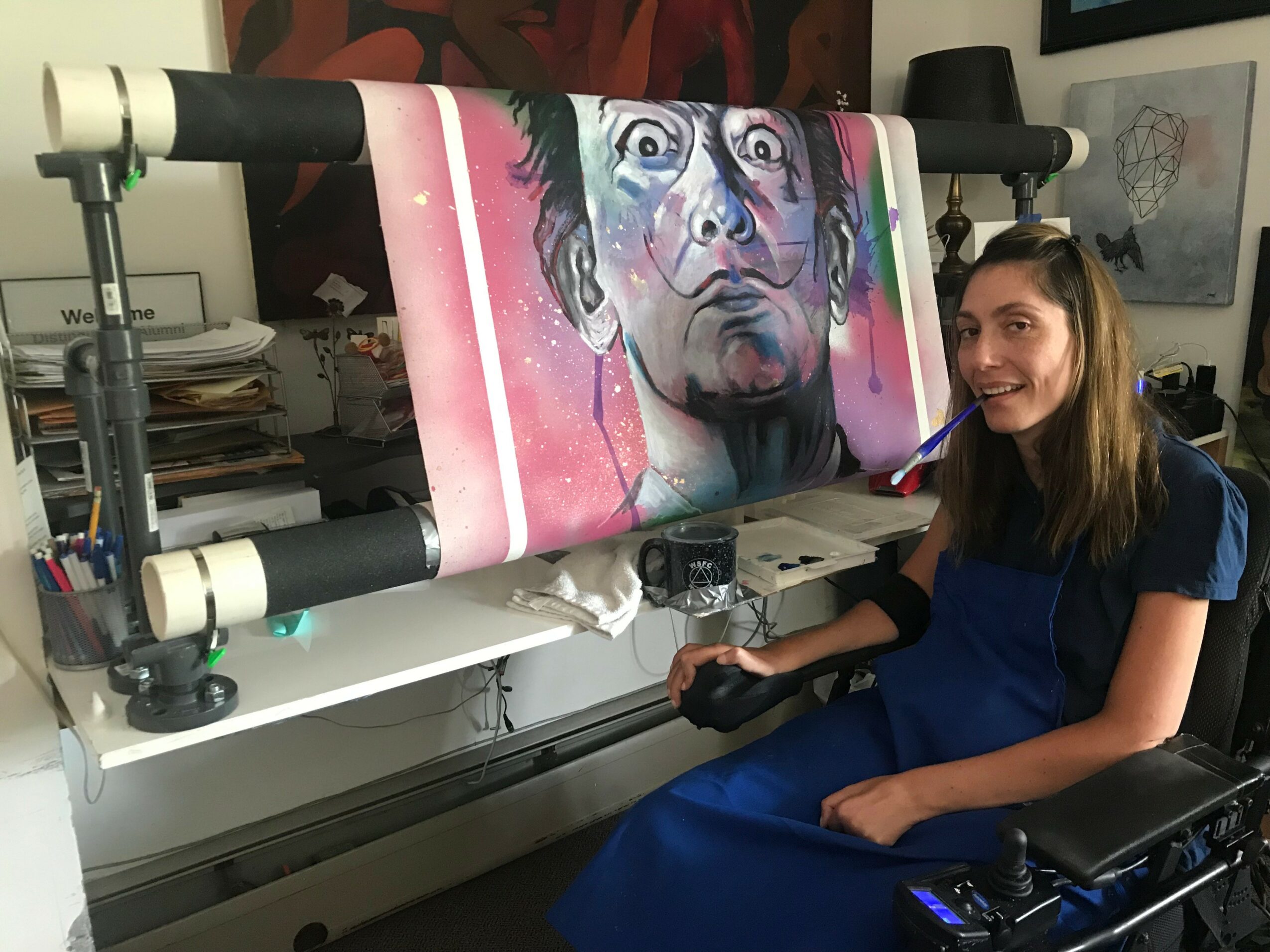 Young woman in a wheelchair smiling next to a large colorful portrait on a printing machine, in a room with artwork and books.