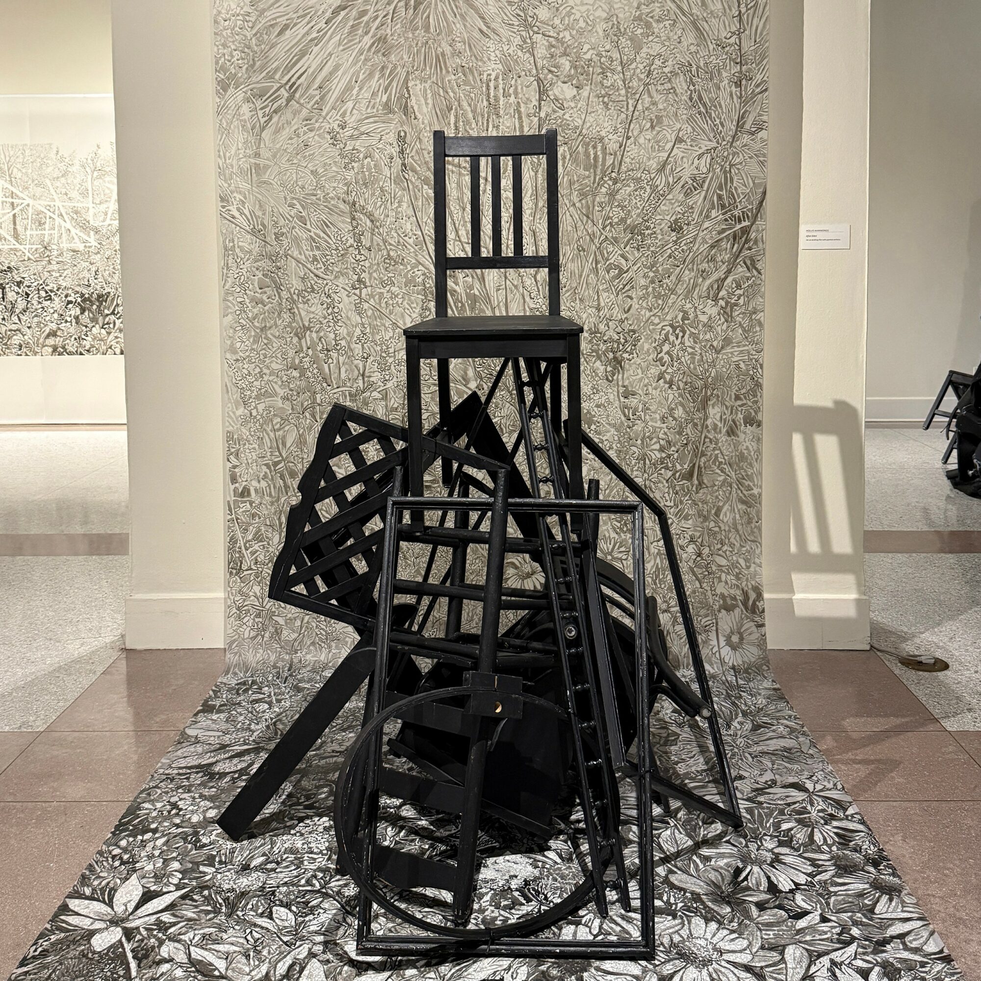 Stacked black chairs and a tall black chair on top, arranged on a patterned rug in an art gallery.