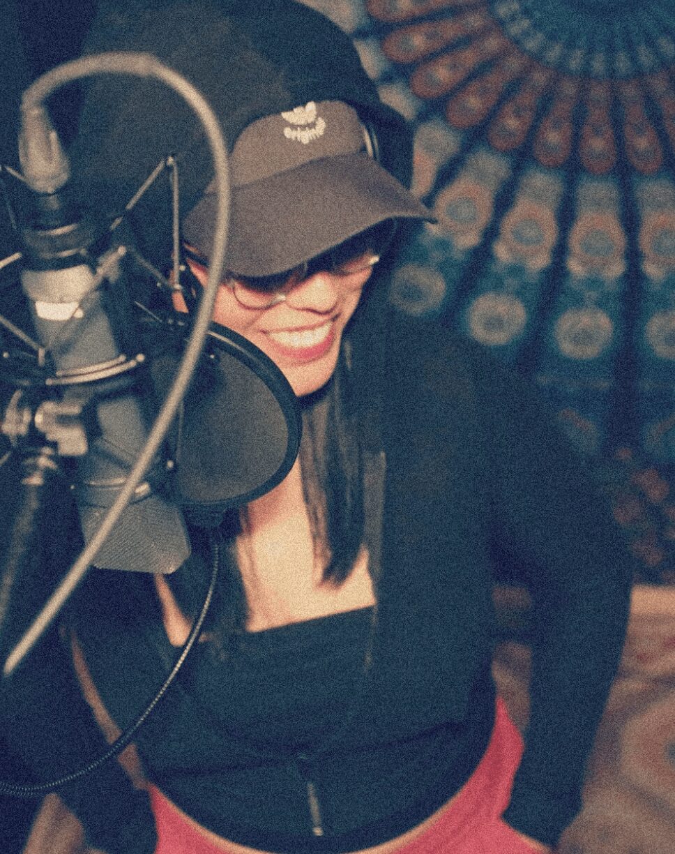 Woman wearing a cap and glasses, smiling, in a recording studio with a microphone and colorful background.
