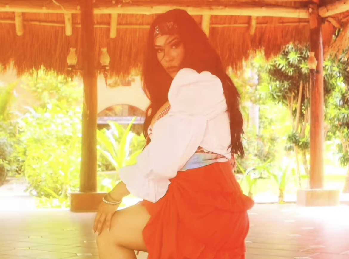 Woman with long dark hair in white blouse and red skirt sitting outdoors under a thatched roof, greenery in background.