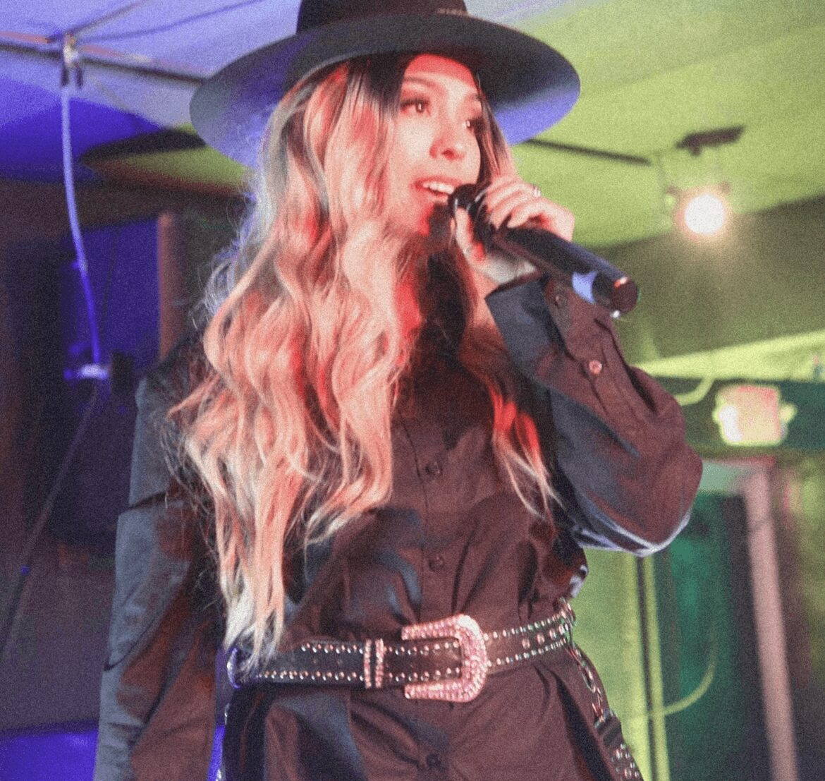Woman singing into a microphone, wearing a wide-brimmed hat and black outfit, with colorful stage lighting.