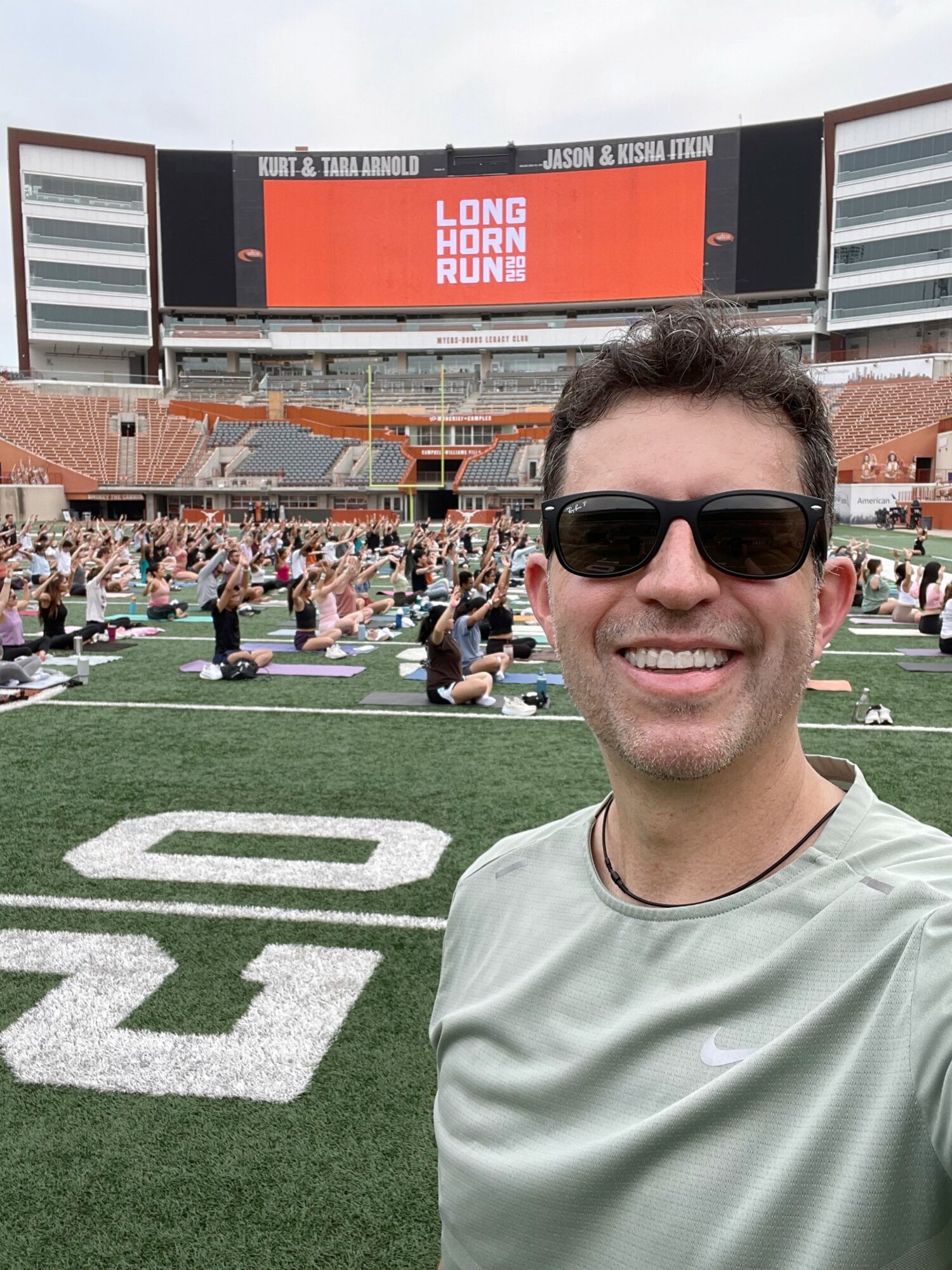 Man wearing sunglasses smiling on a football field with people exercising in the background.