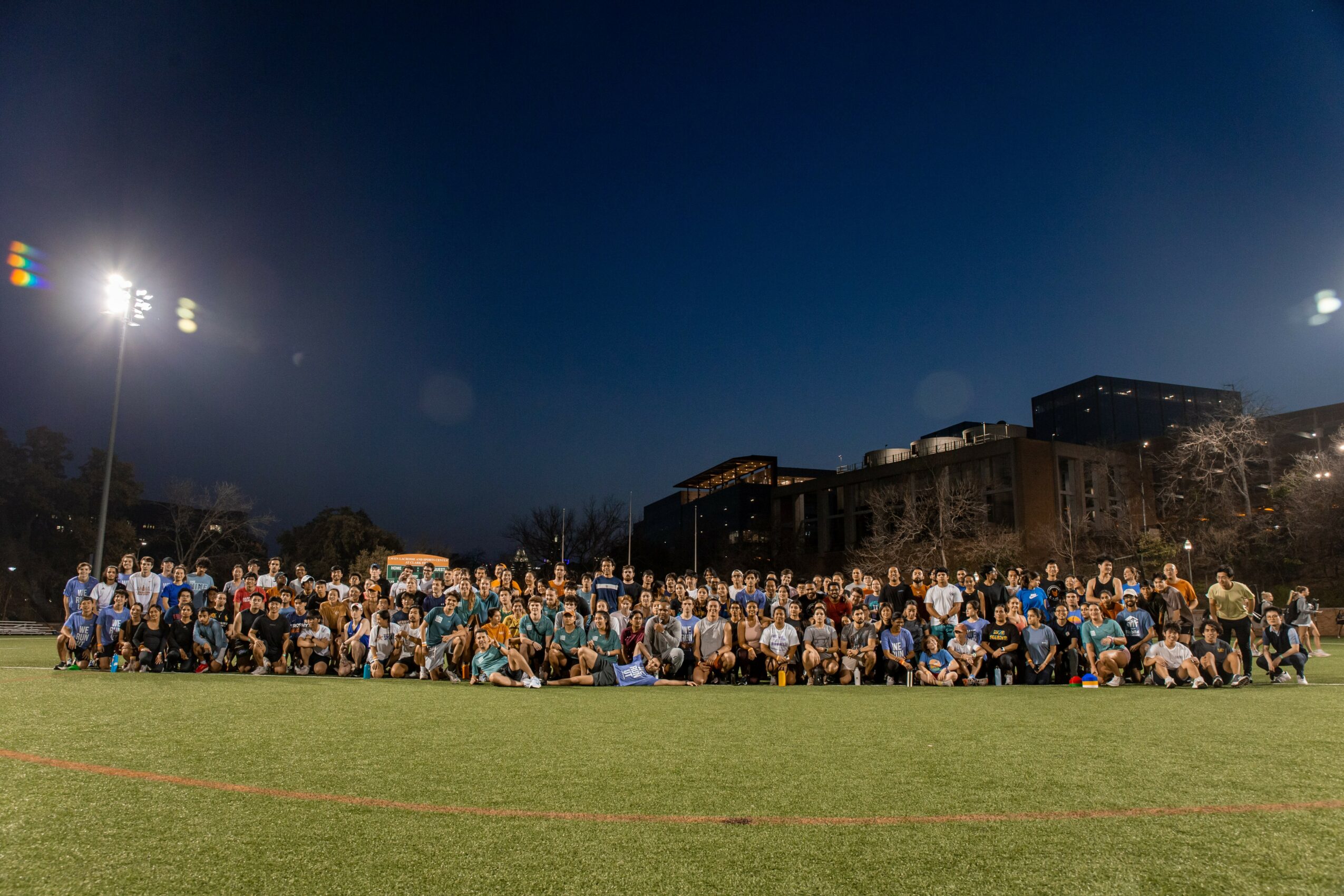 Large group of people gathered on a field at night with buildings and bright lights in the background.