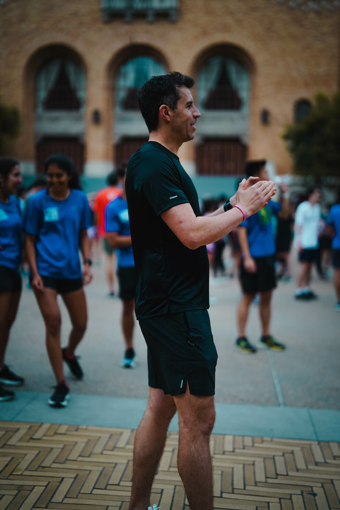 Man clapping outdoors with people in the background, brick building, and trees, during daytime.
