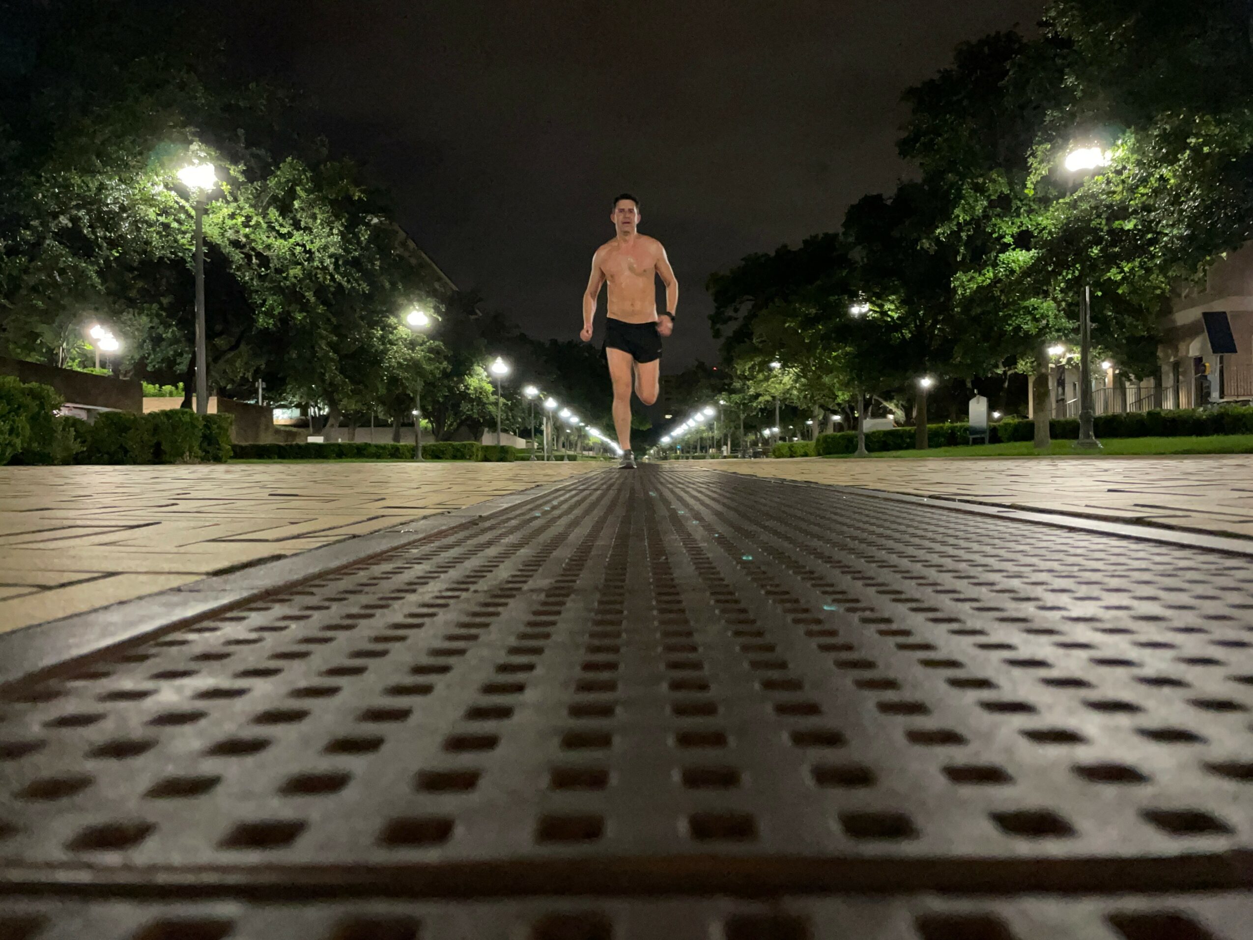 Person walking on a lit outdoor pathway at night, trees and streetlights in the background, low-angle view.