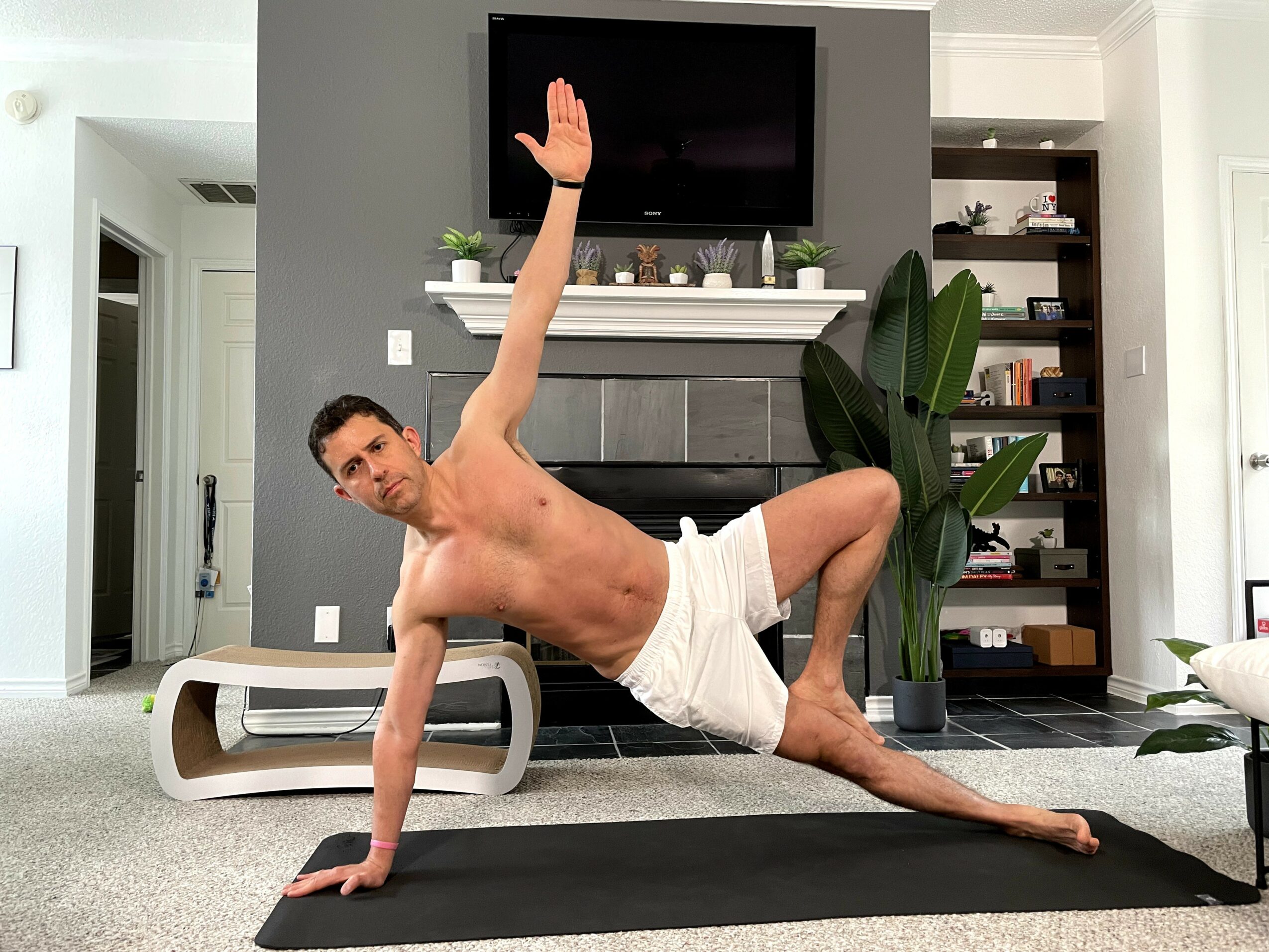 Man performing side plank yoga pose on black mat in living room with fireplace and TV above, plants nearby.