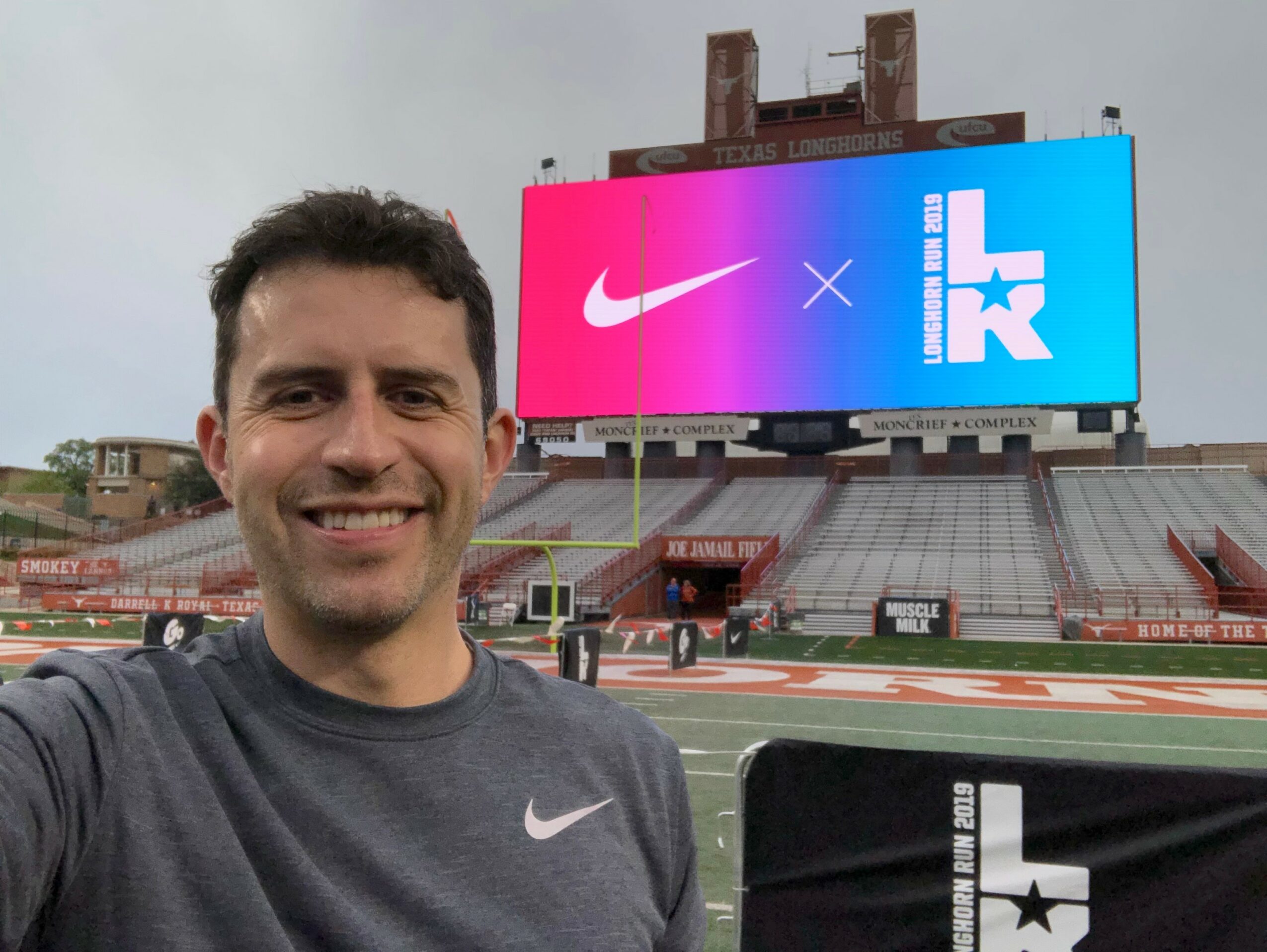 Man smiling in a sports stadium with a large digital screen behind him displaying Nike and other logos.