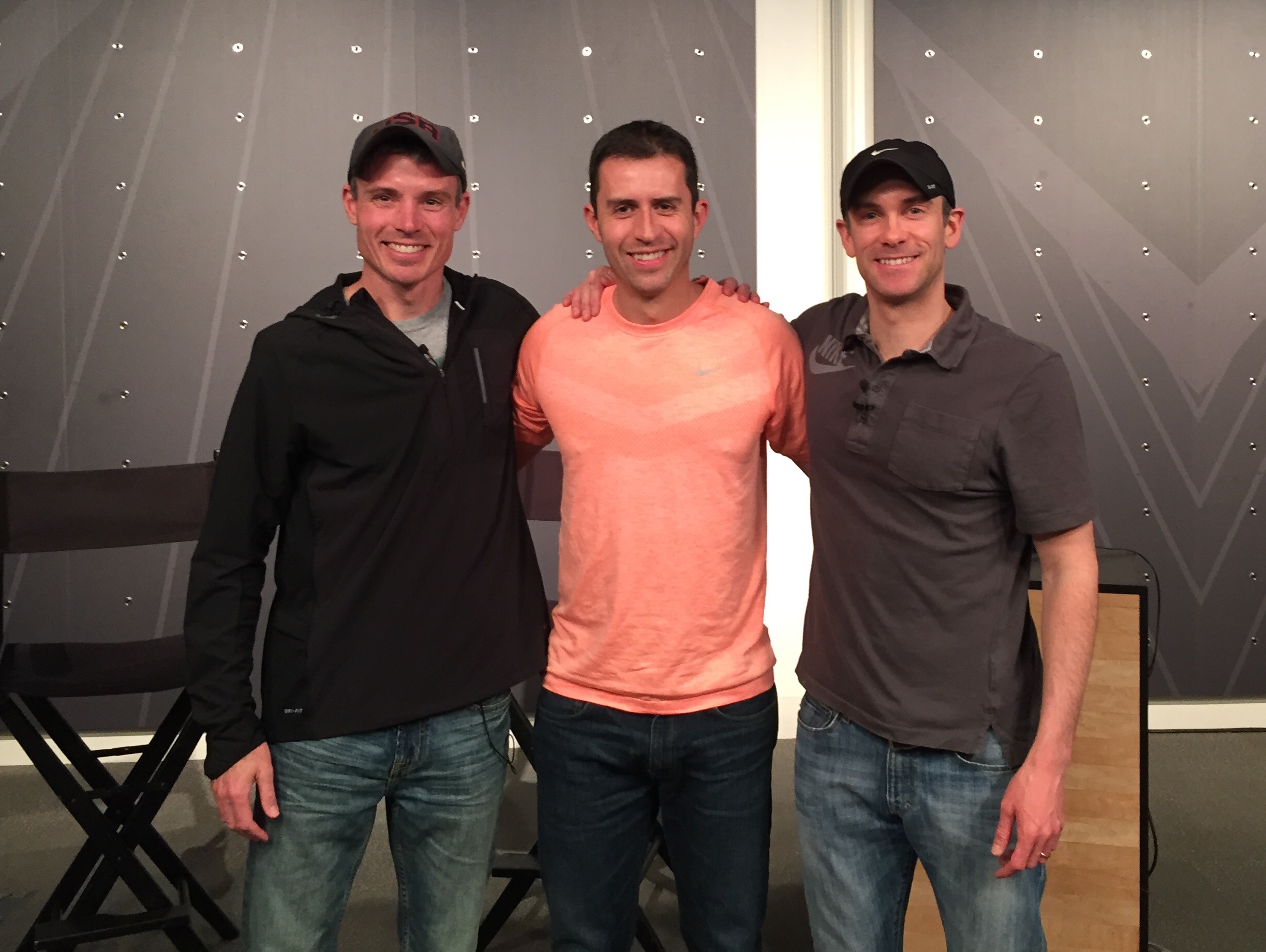 Three men stand together indoors, smiling, with a patterned wall behind them.