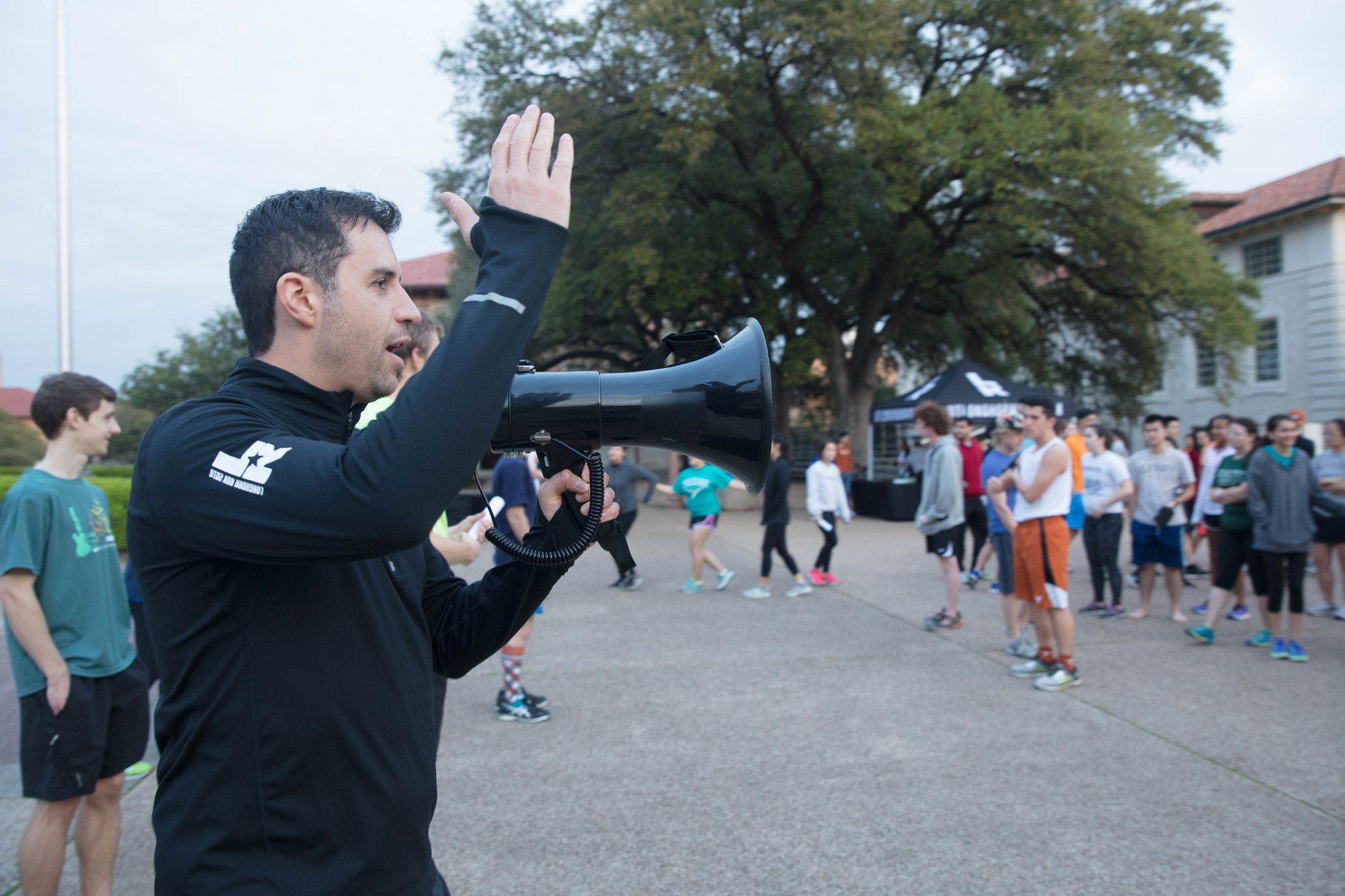 Man with a megaphone speaking to a group outdoors, trees and buildings in background, people gathered around.