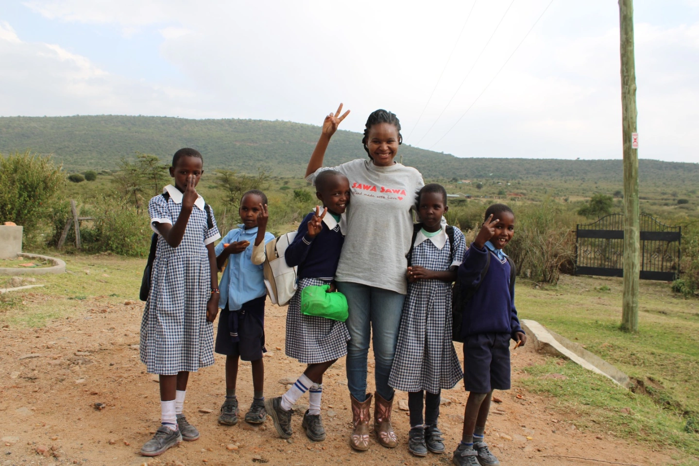 Group of children and a woman standing outdoors in a rural area, some children in school uniforms, woman making peace sign.