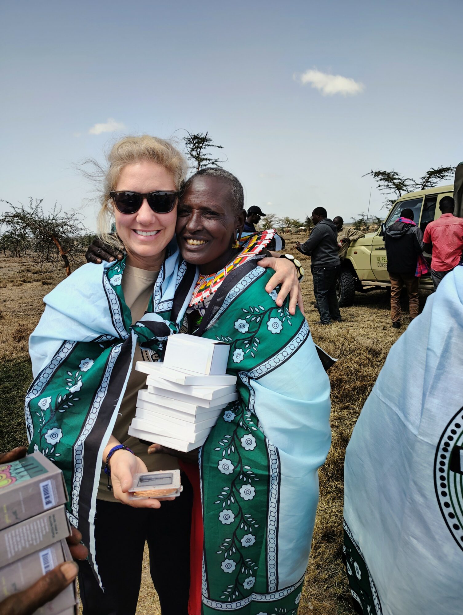 Two women hugging outdoors, one holding a stack of white boxes and a phone, smiling, with trees and vehicles in background.