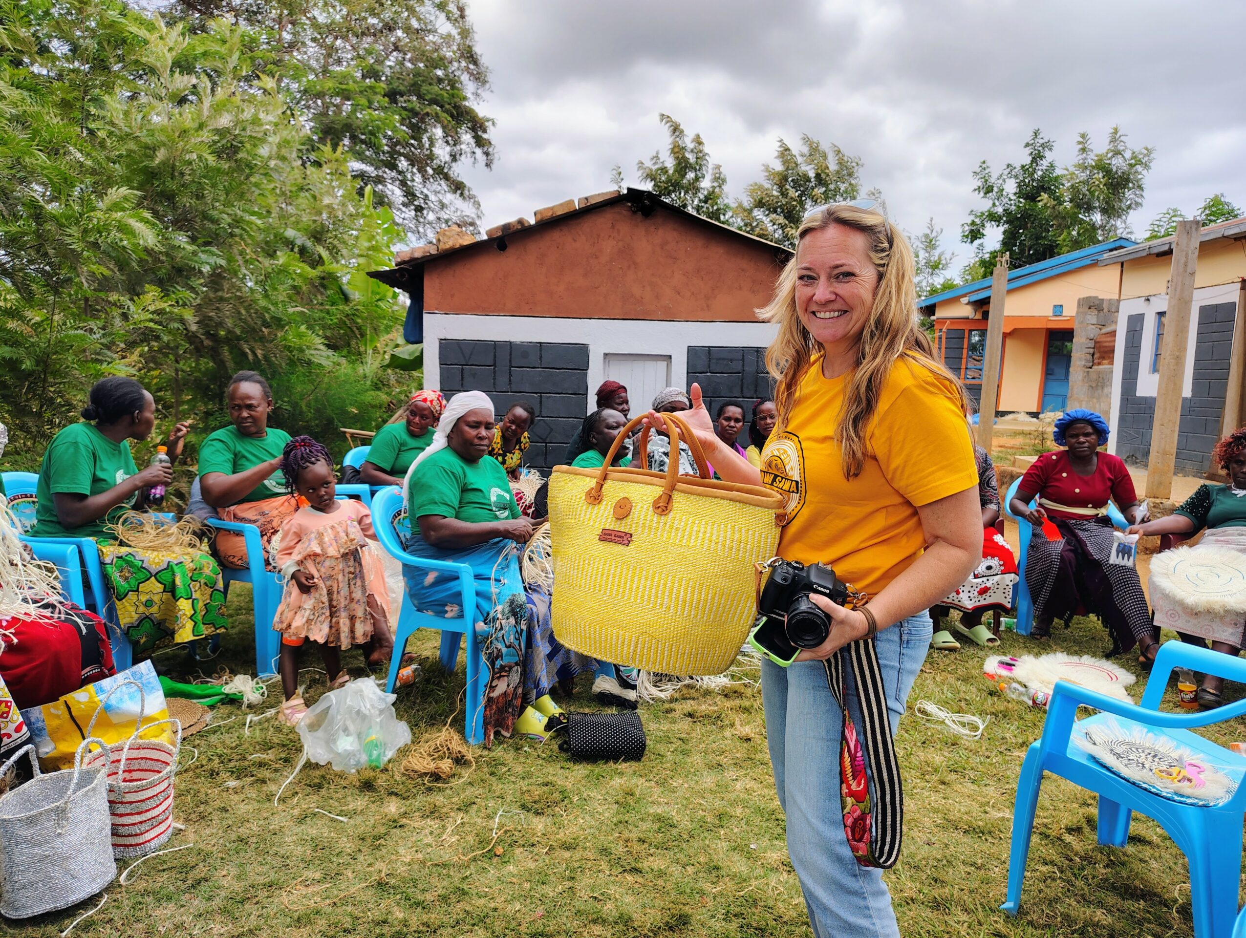 Woman in yellow shirt holding a yellow bag and camera, group of people sitting outdoors on blue chairs, buildings in background.