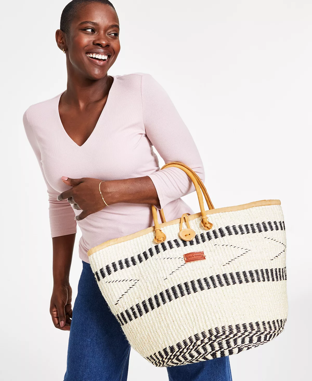 Smiling woman holding a large woven bag with black and white patterns, wearing a light pink top and blue jeans.