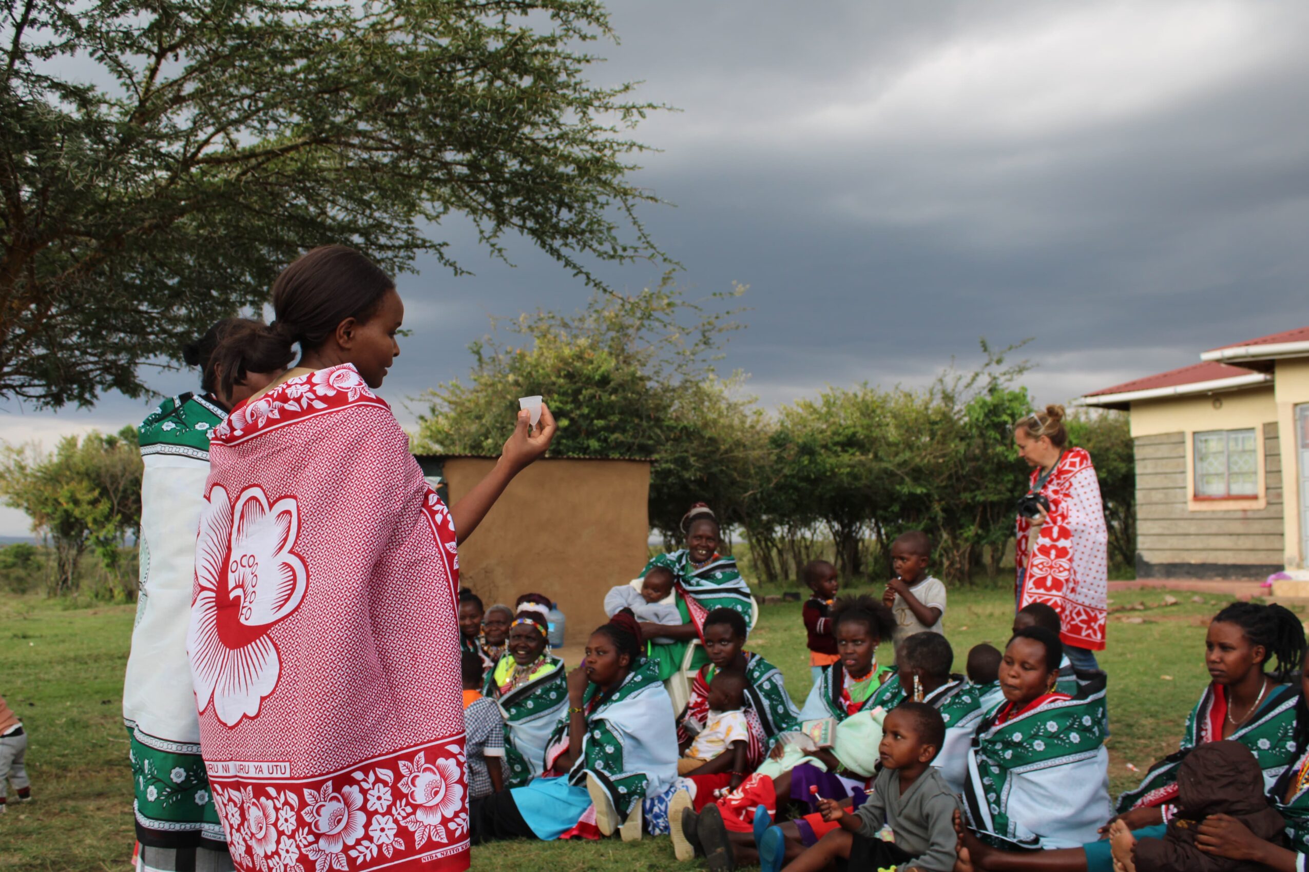 Group of children and adults outdoors, some children wearing school uniforms, under cloudy sky, trees, and buildings in background.