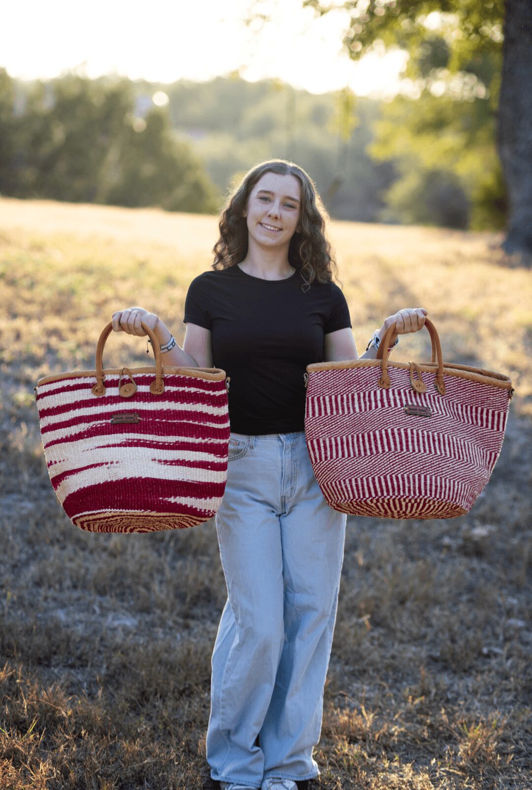 Young woman holding two large striped bags outdoors in a grassy area with trees in the background.