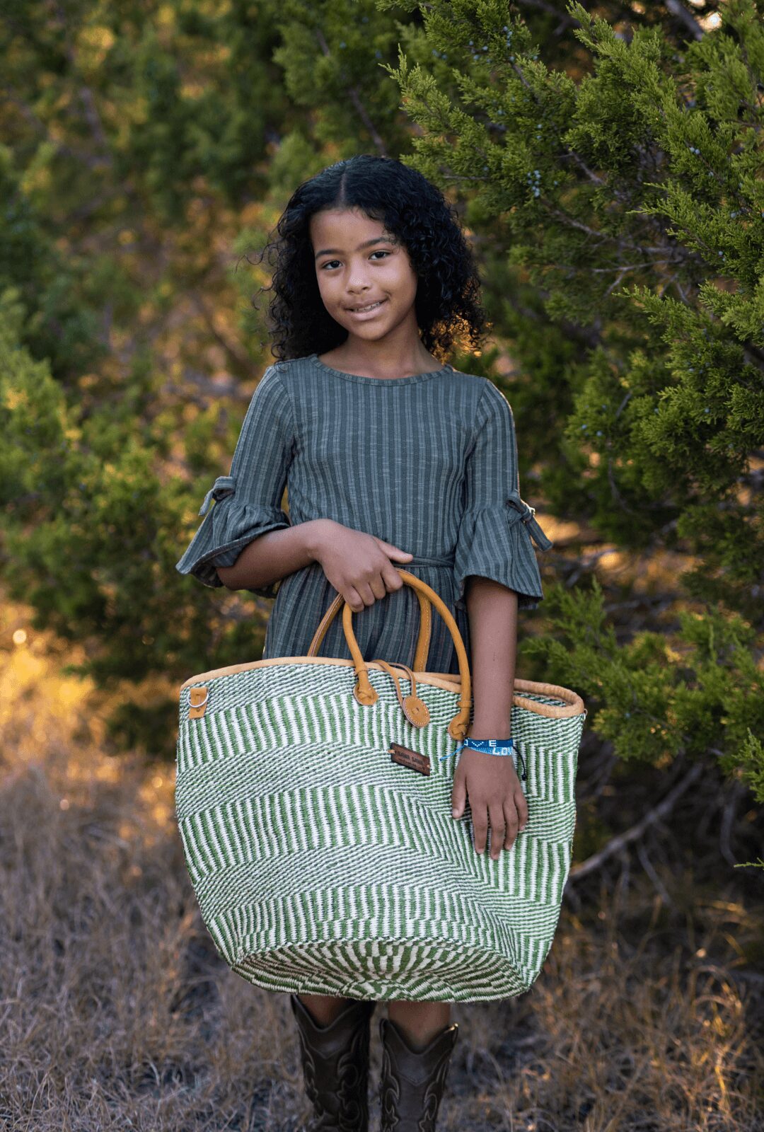 Young girl with curly hair holding a large striped bag outdoors, surrounded by trees and greenery.