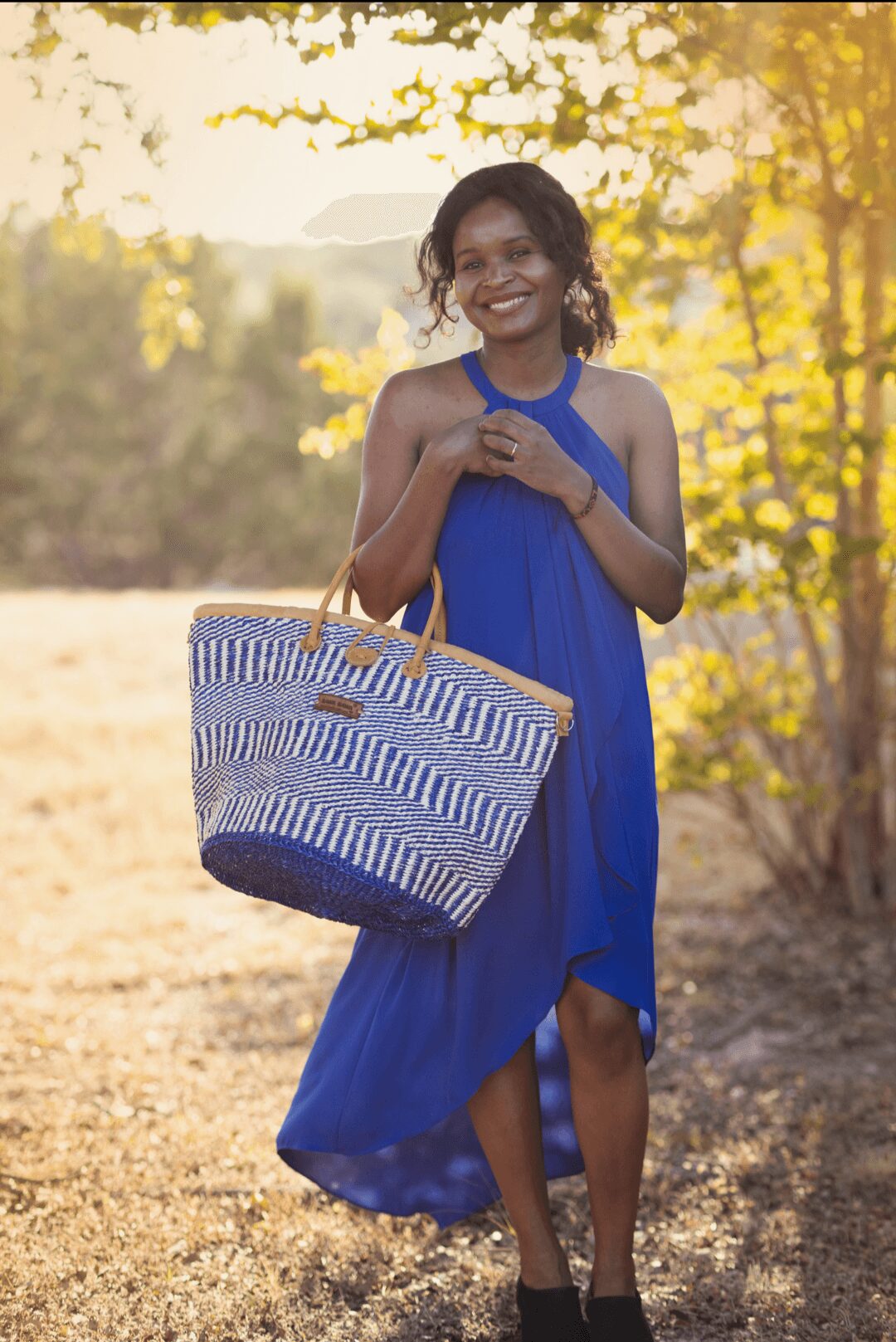 Smiling woman in a blue dress holding a large striped bag outdoors with trees and sunlight in background.