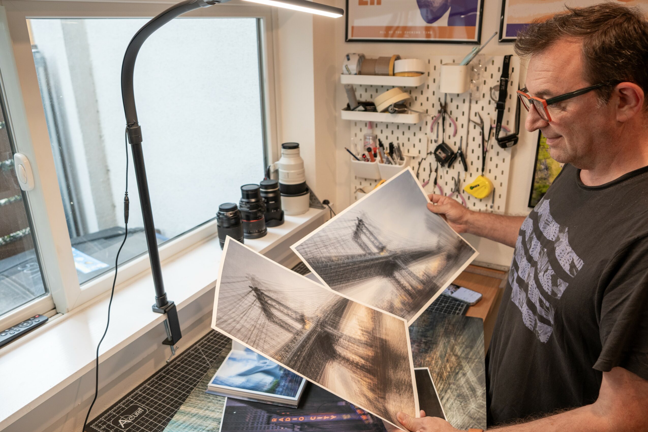 Man examining photographs on a workbench in a workshop, with tools and supplies on the wall behind him.