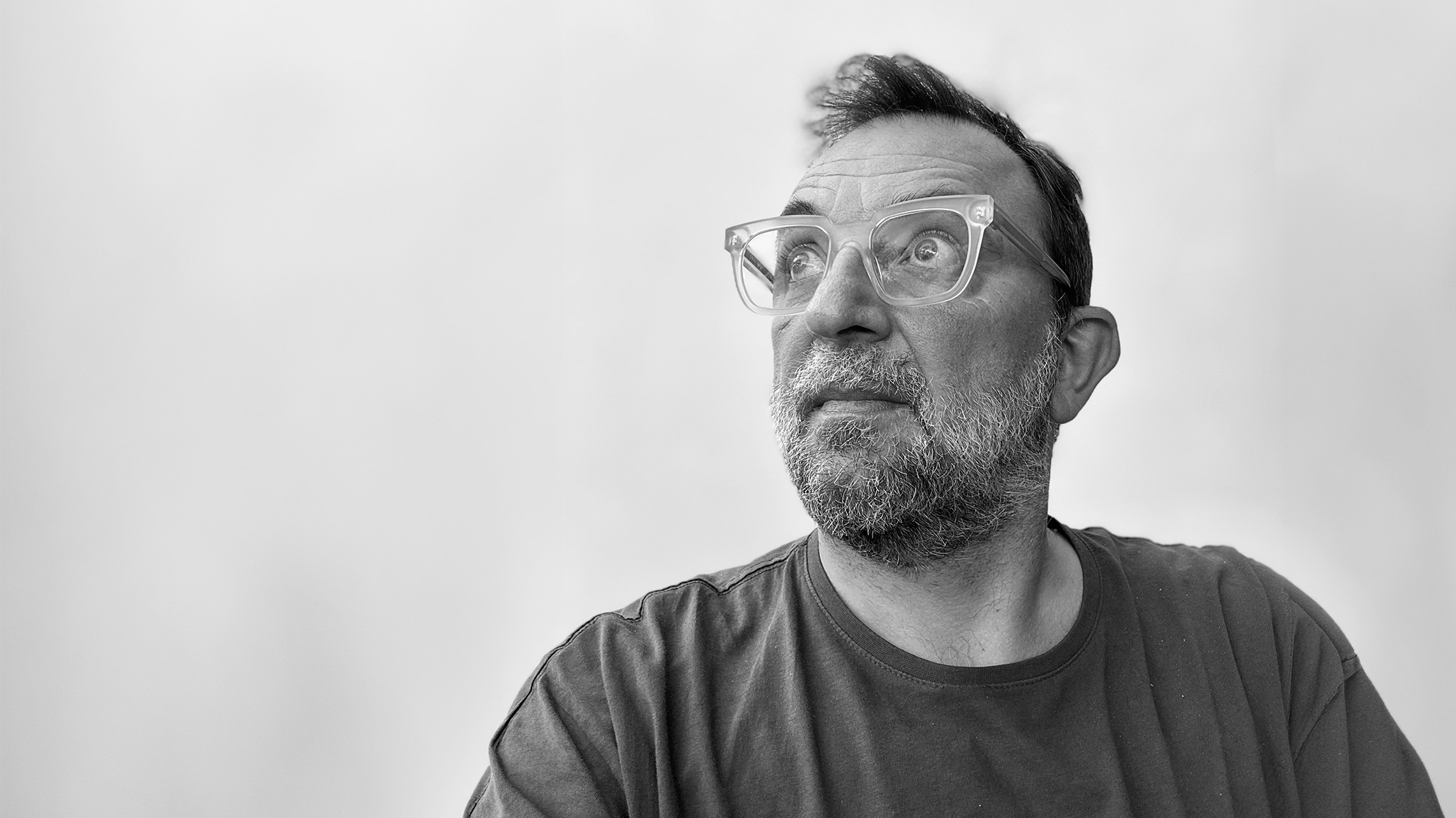 A man with glasses and a beard looks to the side against a plain background.