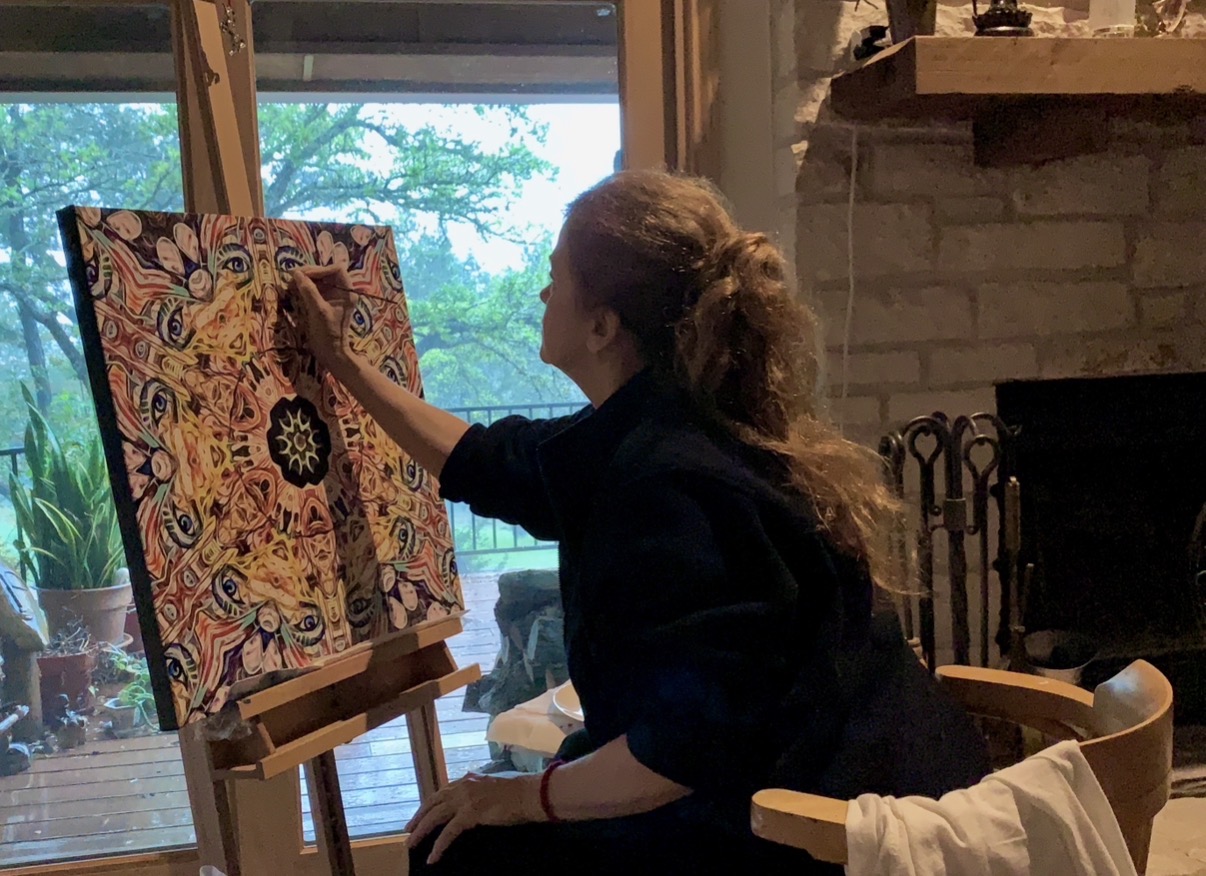 Person painting on canvas indoors, sitting near a window with trees outside, using an easel, with a fireplace nearby.