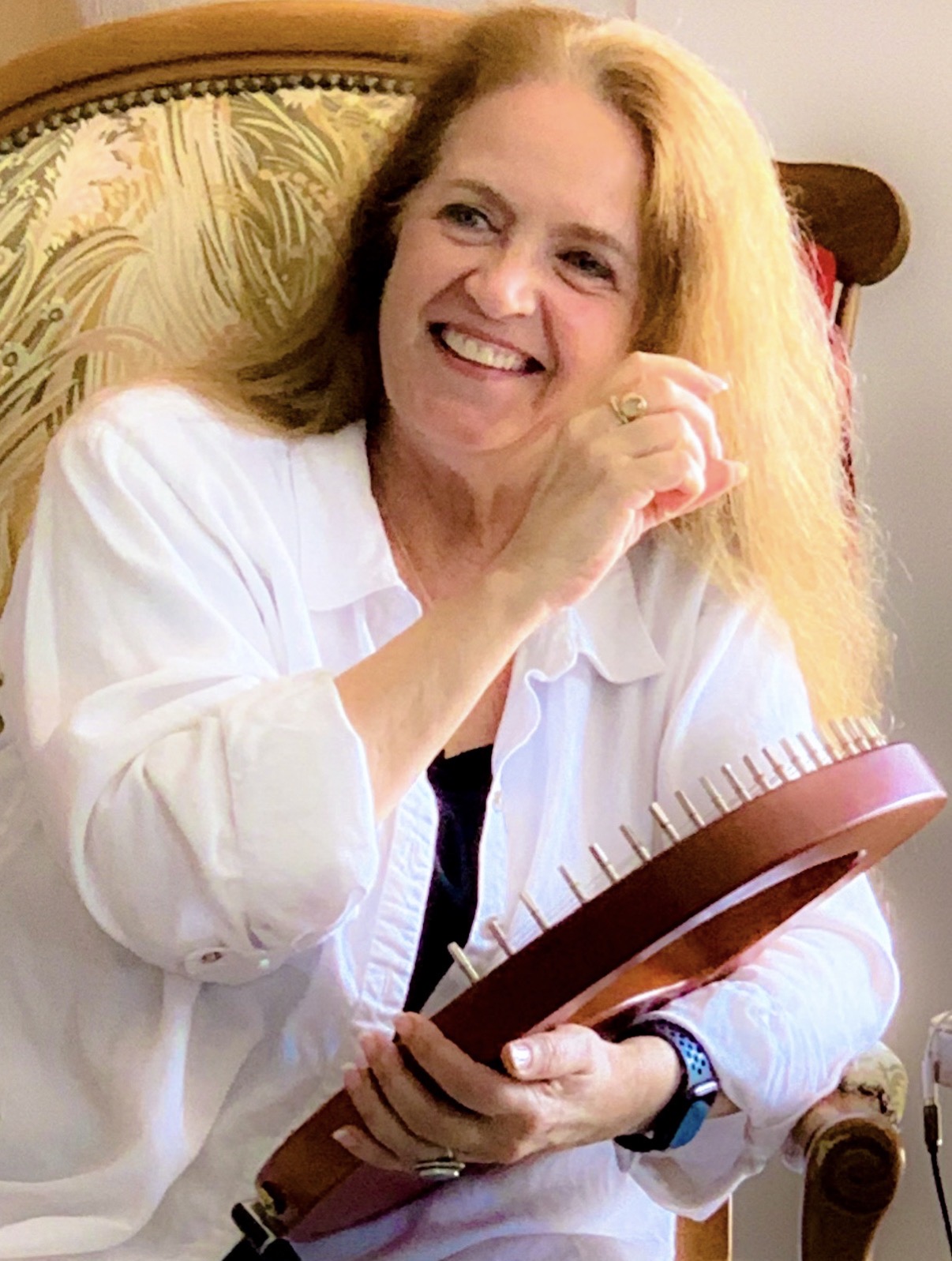 Smiling woman with long blonde hair, wearing a white shirt, sitting on a chair, holding a brown object.