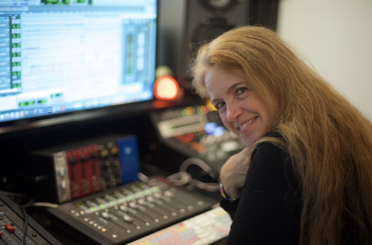 Woman with long hair smiling at a mixing console in a recording studio, computer screen in background