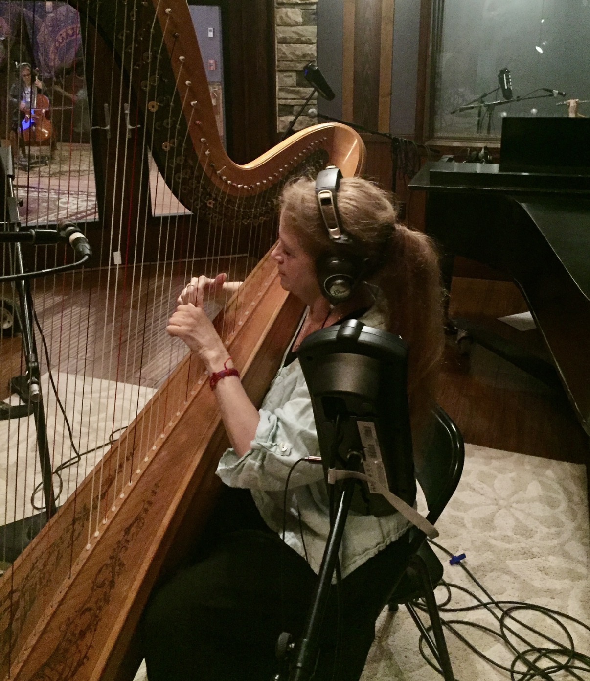 Young woman with headphones playing a harp in a recording studio, sitting at a wooden harp with a window behind her.