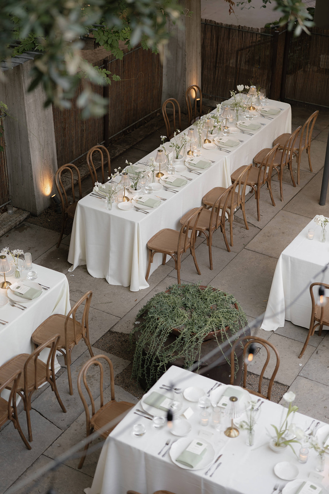 Long dining table with white tablecloth, surrounded by wooden chairs, set with plates, glasses, and cutlery, in an outdoor setting with greenery.