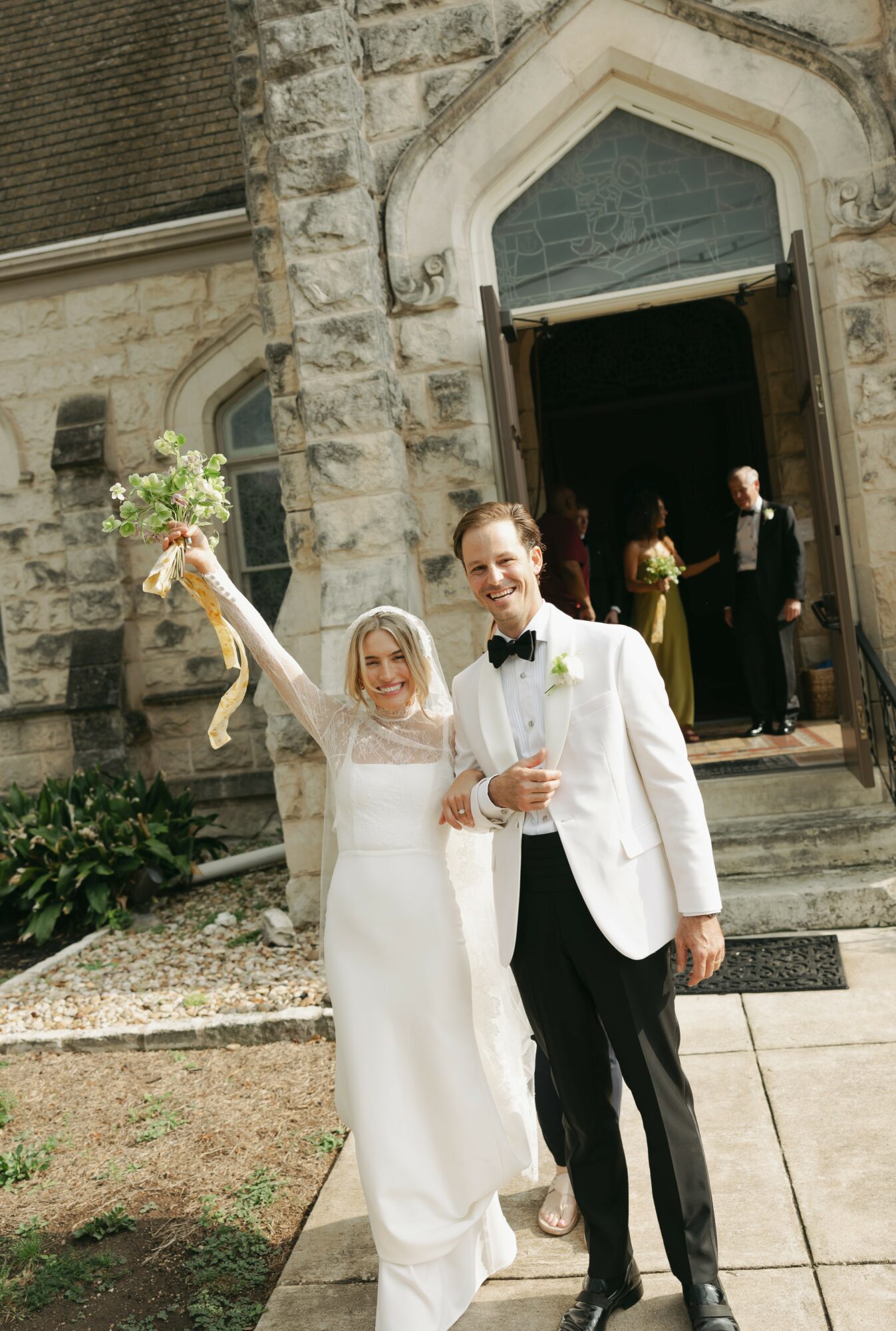 Bride and groom celebrating outside a stone church, bride holding a bouquet and raising her arm, groom smiling with a glass.