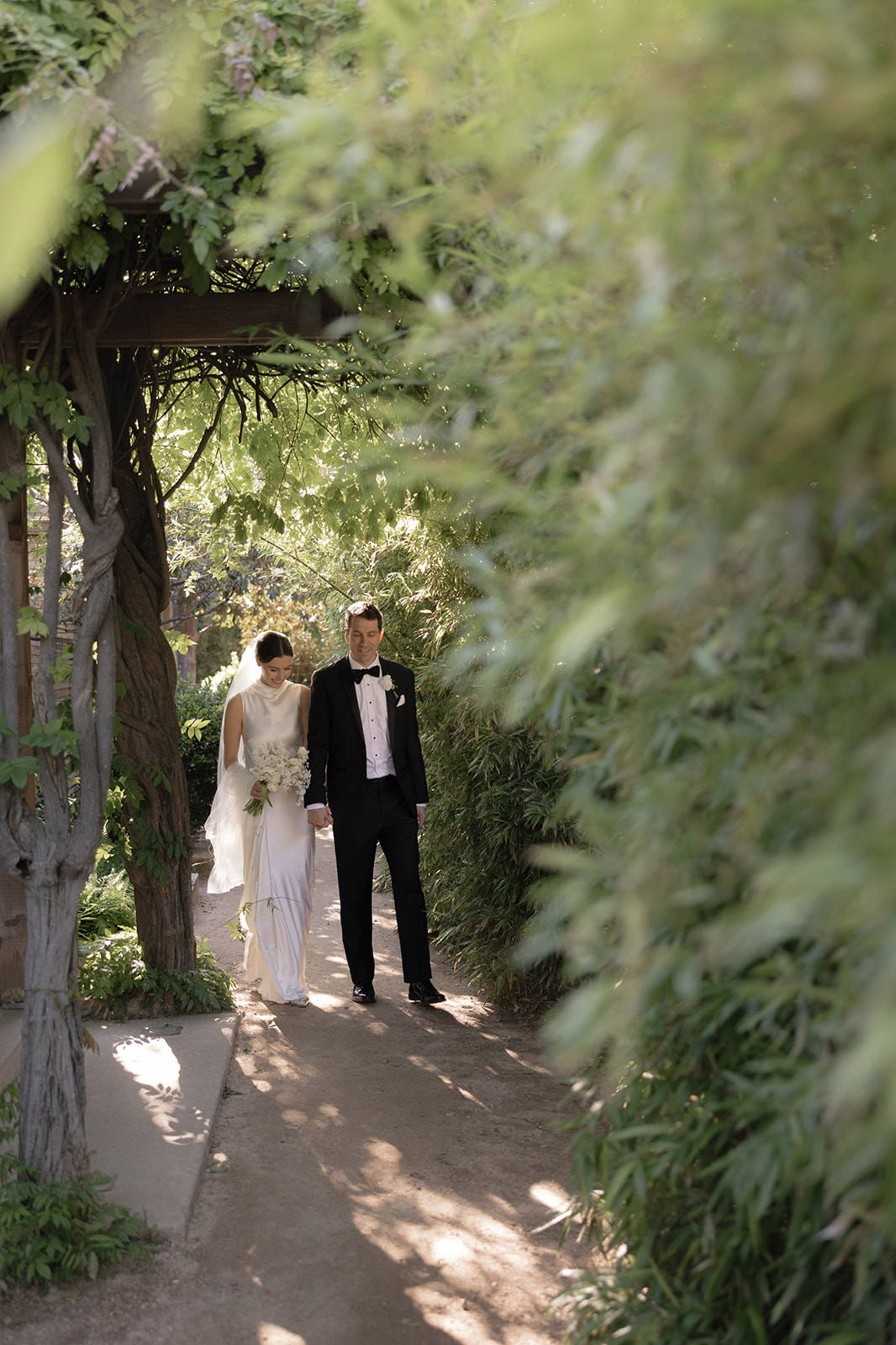 A couple dressed in formal attire walking on a shaded garden path surrounded by greenery.