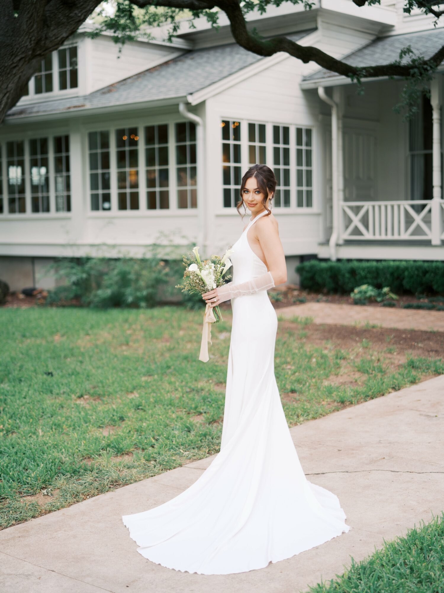 Bride in a white wedding dress holding a bouquet, standing on a sidewalk in front of a house with large windows.