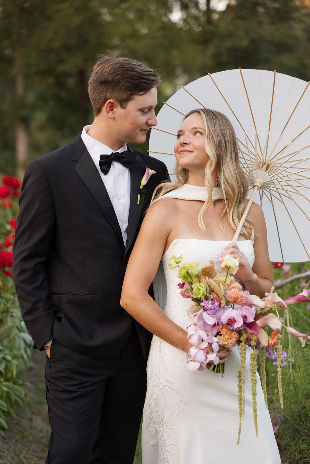 A couple dressed in formal attire standing outdoors with a white parasol, surrounded by greenery and flowers, holding a bouquet.