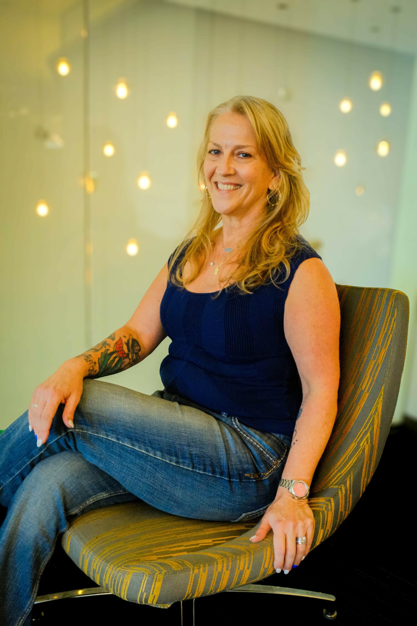 Woman with blonde hair smiling, sitting on a striped chair, wearing a dark sleeveless top and jeans, in a decorated room.