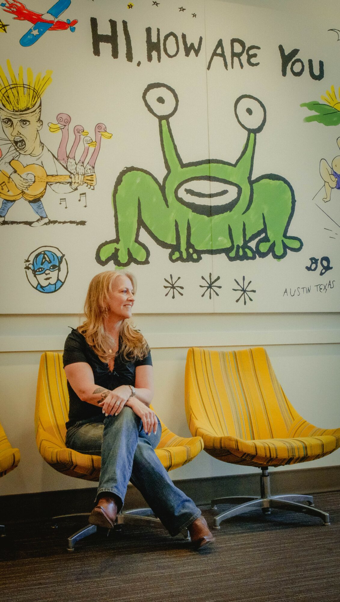 Woman sitting on yellow chair with colorful wall art behind her, including a green cartoon alien and a person with a crown.