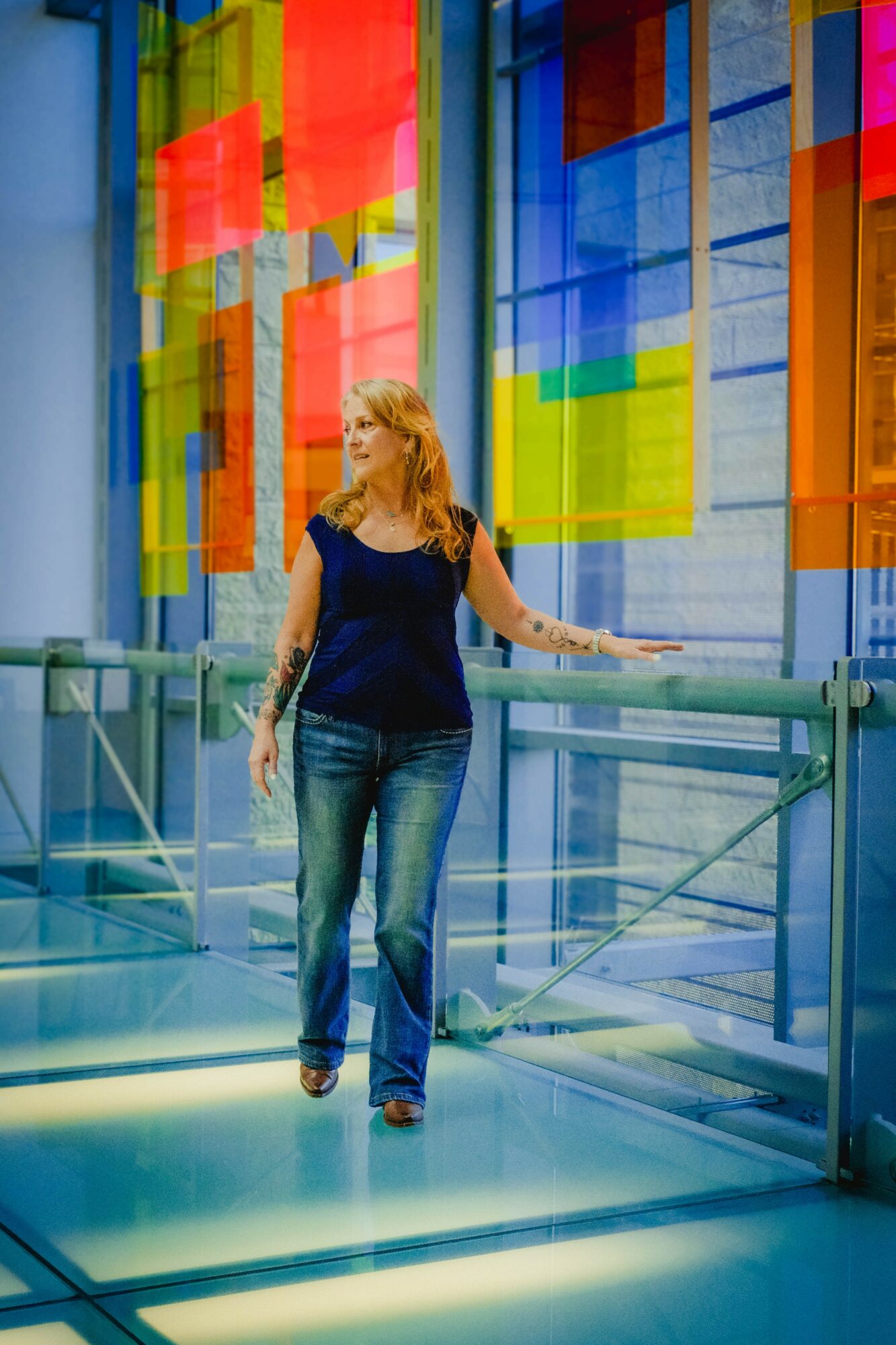 Woman with blonde hair in a navy top and jeans walking on a glass bridge in front of colorful glass panels.