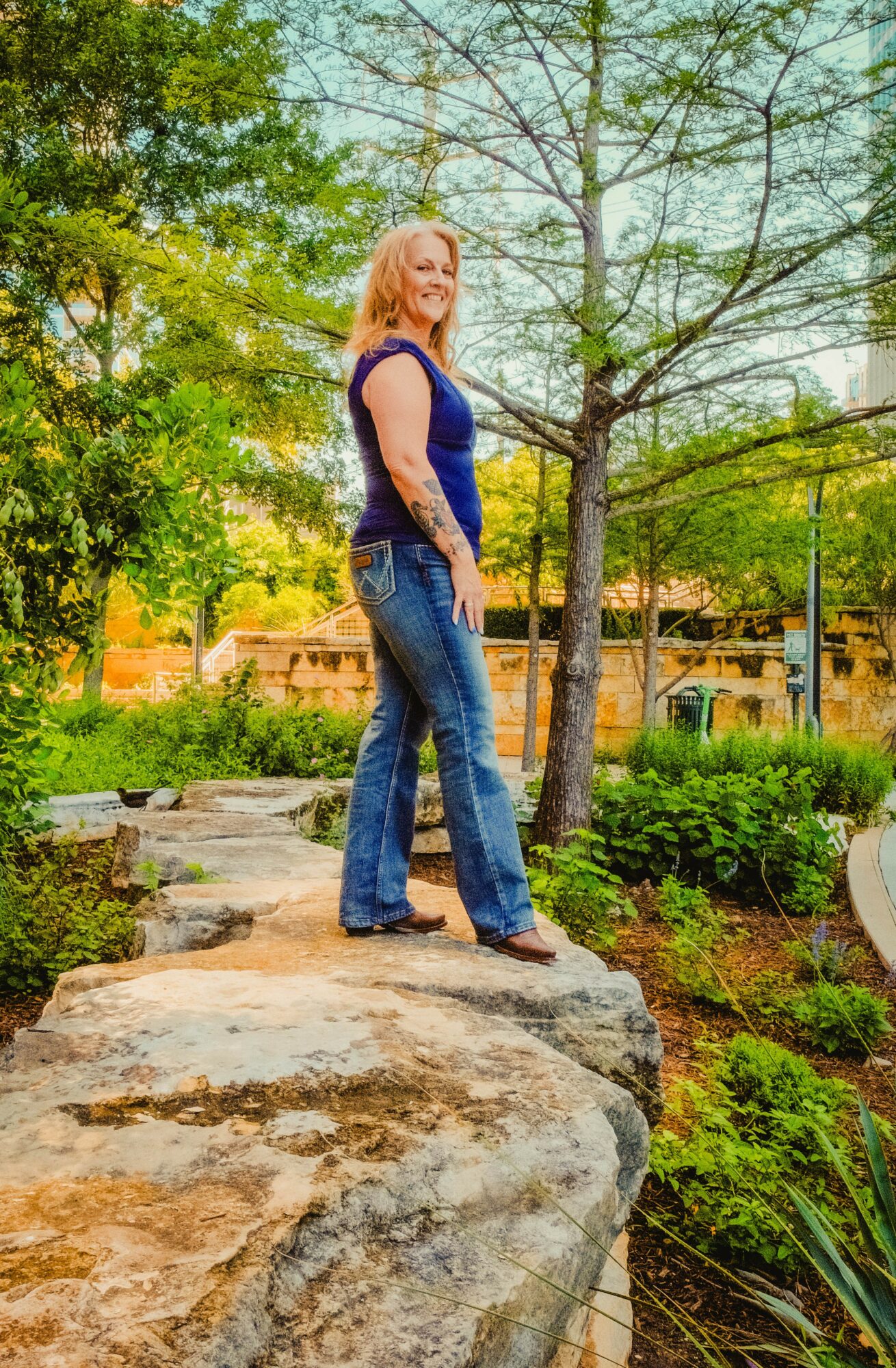 Woman standing on a large rock in a park with trees and greenery, smiling, wearing a blue top and jeans.