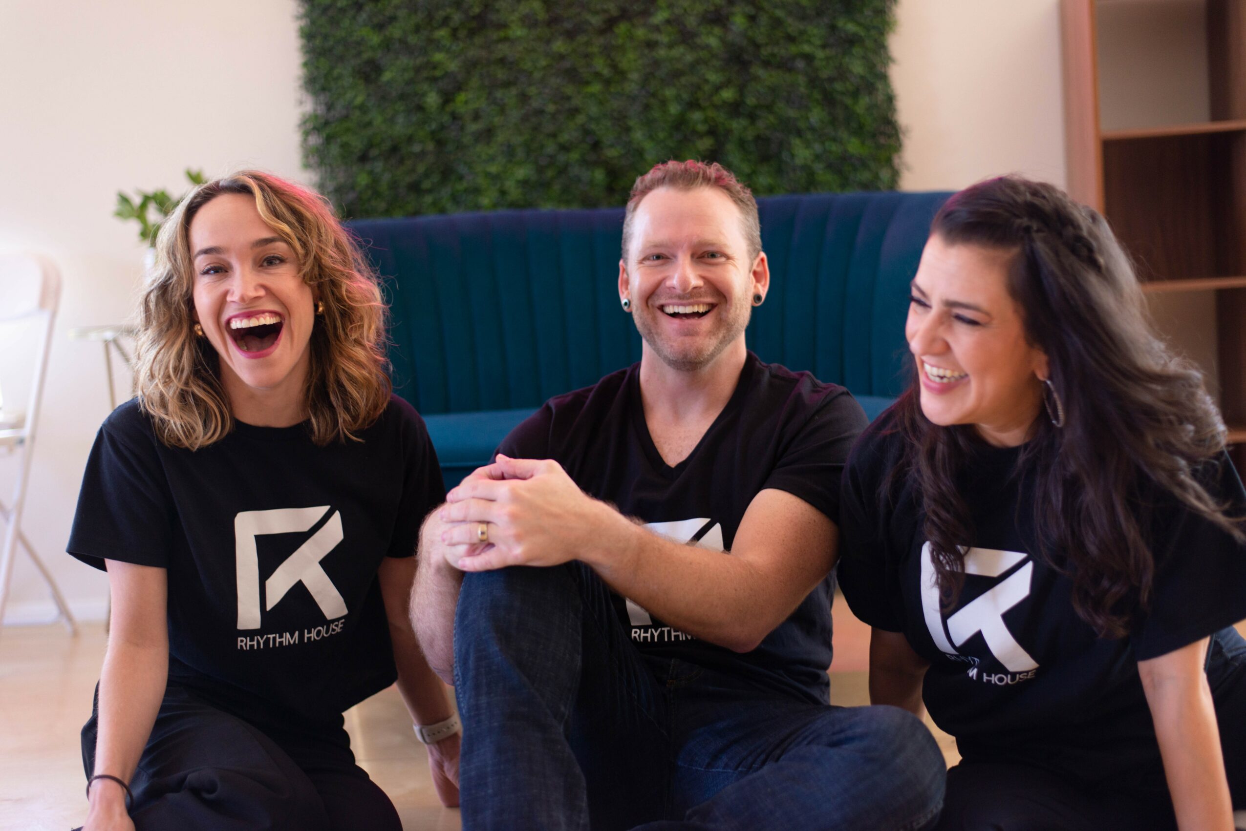 Three people sitting on a couch, smiling and laughing, wearing matching black T-shirts with a logo.