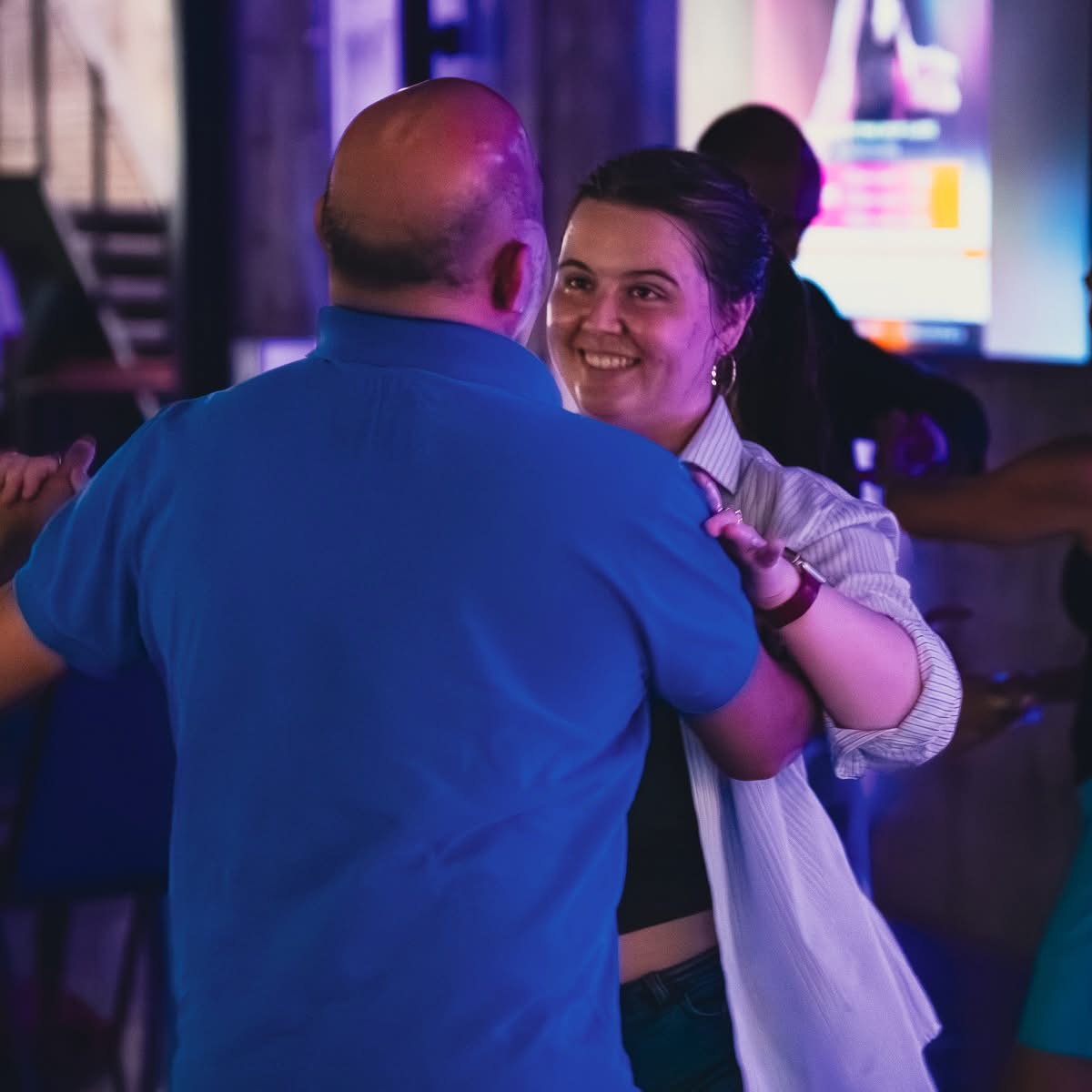 Two people dancing, smiling, in a dimly lit indoor setting with colorful lights.